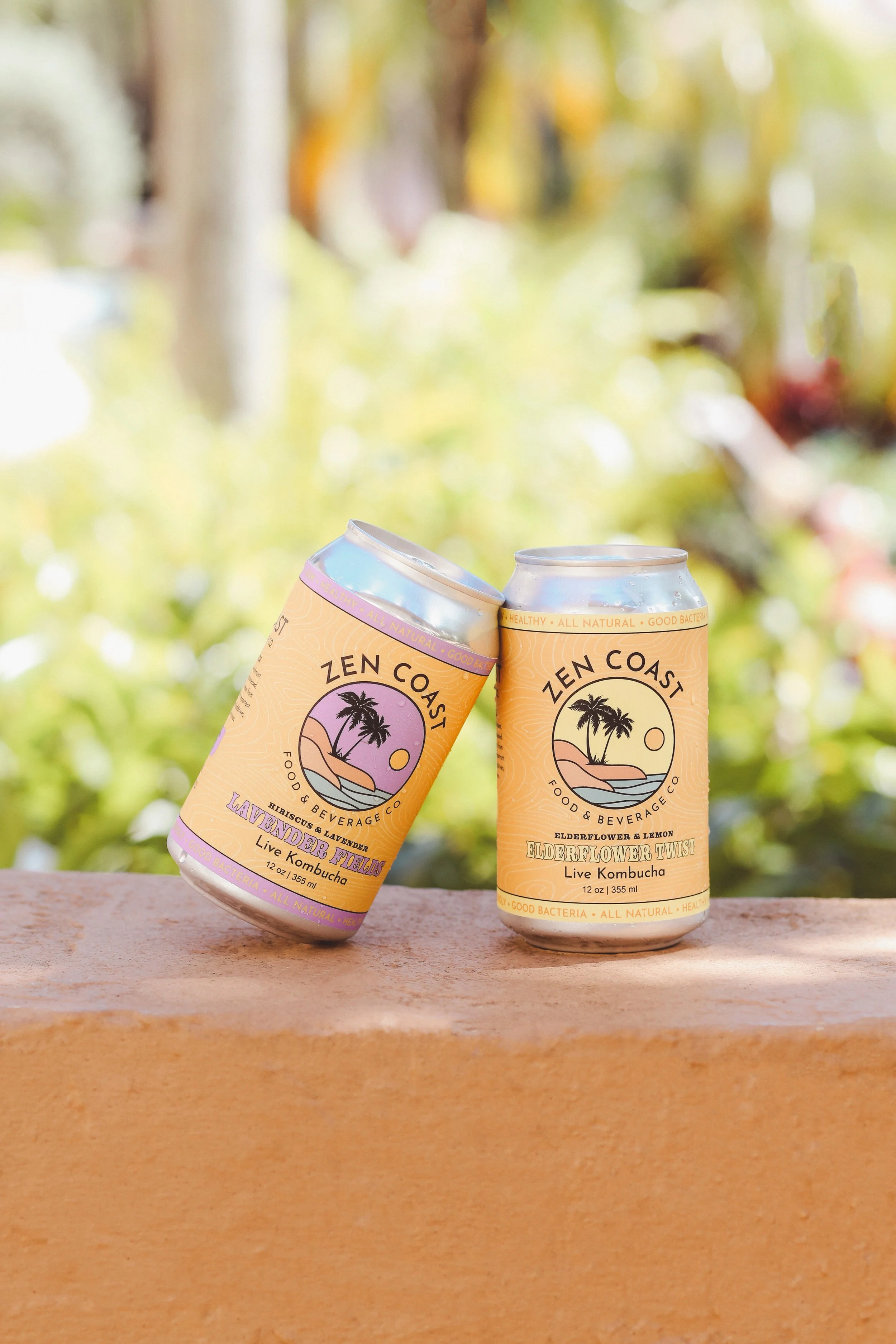 Two cans of Zen Coast kombucha on a wooden surface with a blurred green and yellow foliage background. One can is labeled Lavender Fields, and the other is Elderflower Twist.