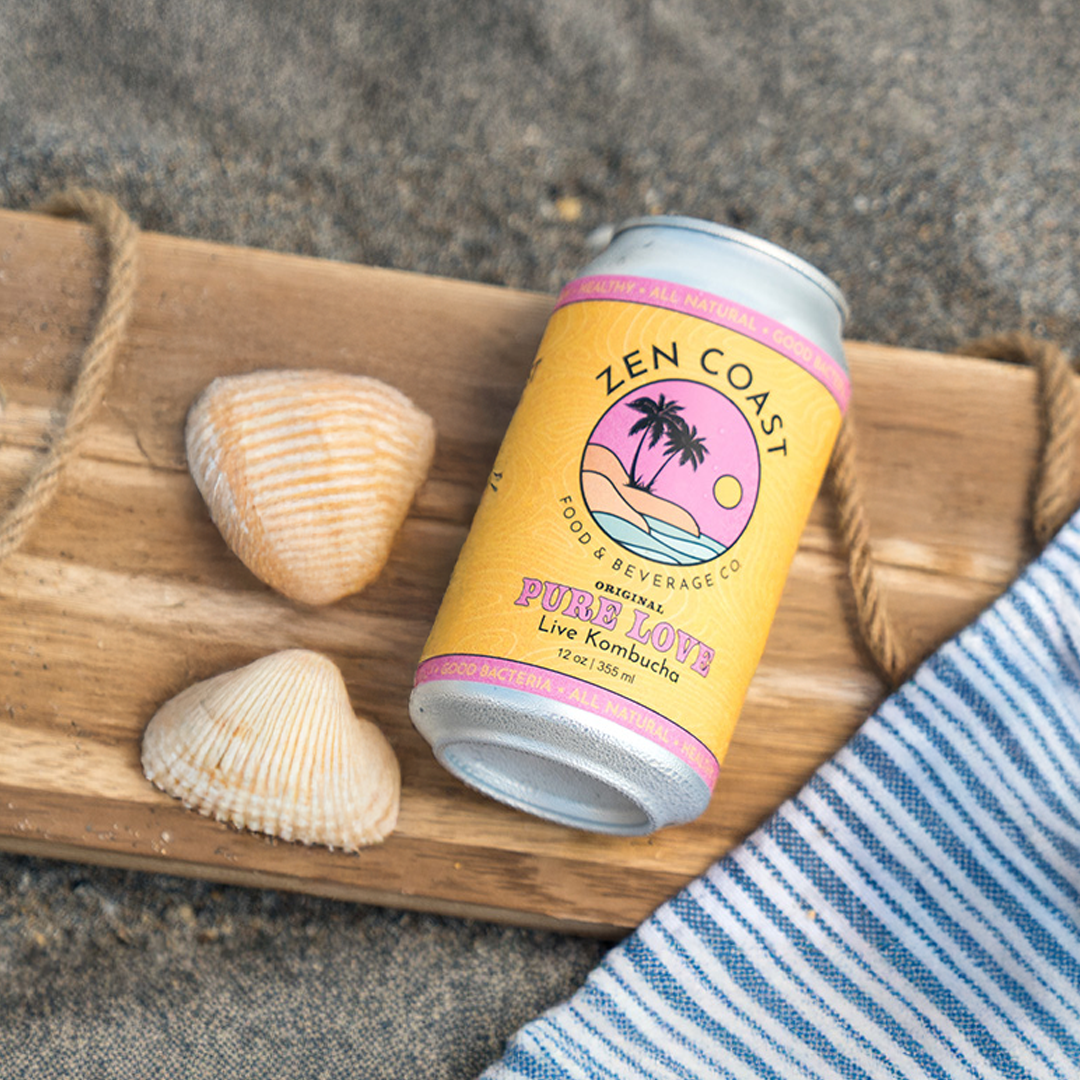 A can of Zen Coast Pure Love kombucha placed on a wooden tray on sandy ground, with two seashells and a blue striped cloth nearby.