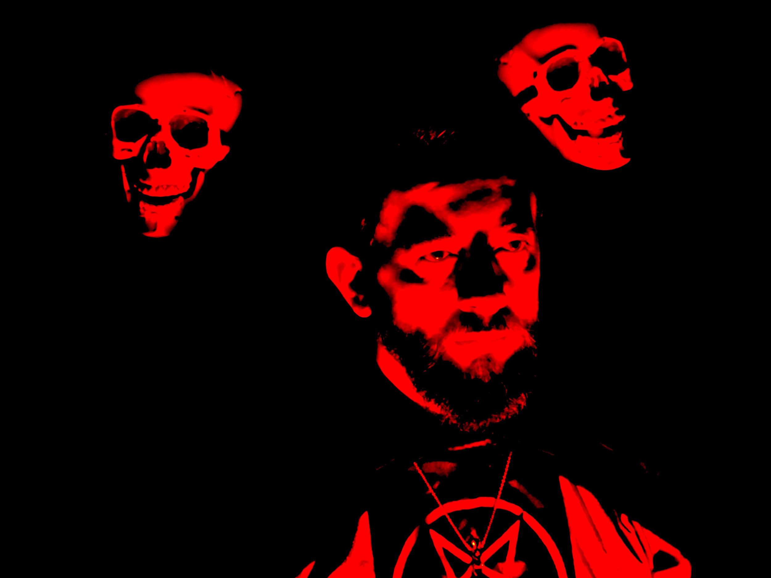 A dark image featuring a person with a serious expression, illuminated in red light, wearing a pendant. Two red skulls are positioned near the person's head, also highlighted in red against a black background.