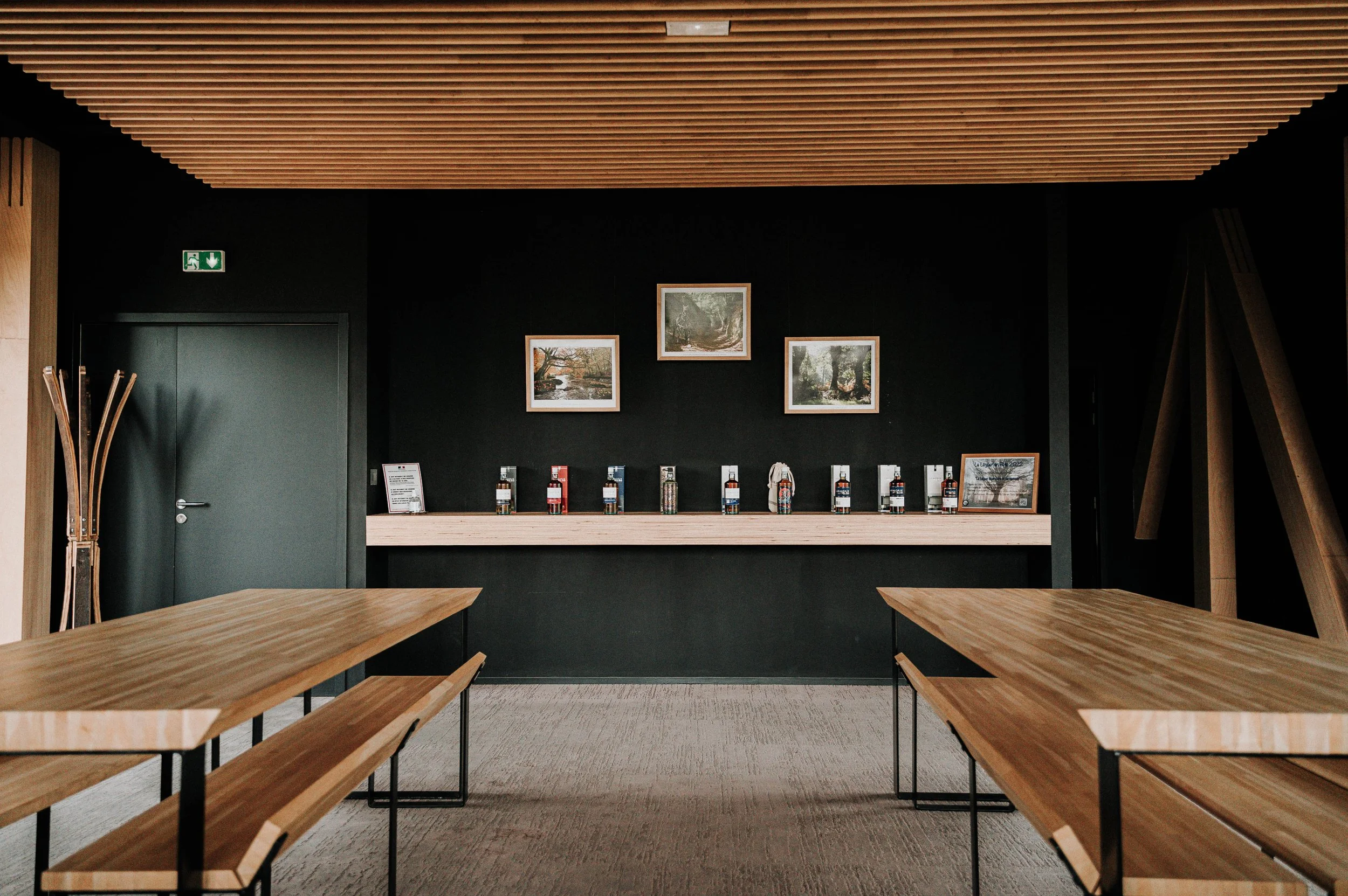 Salle de réunion avec tables en bois, mur noir avec trois tableaux et une rangée de bouteilles derrière une étagère en bois.