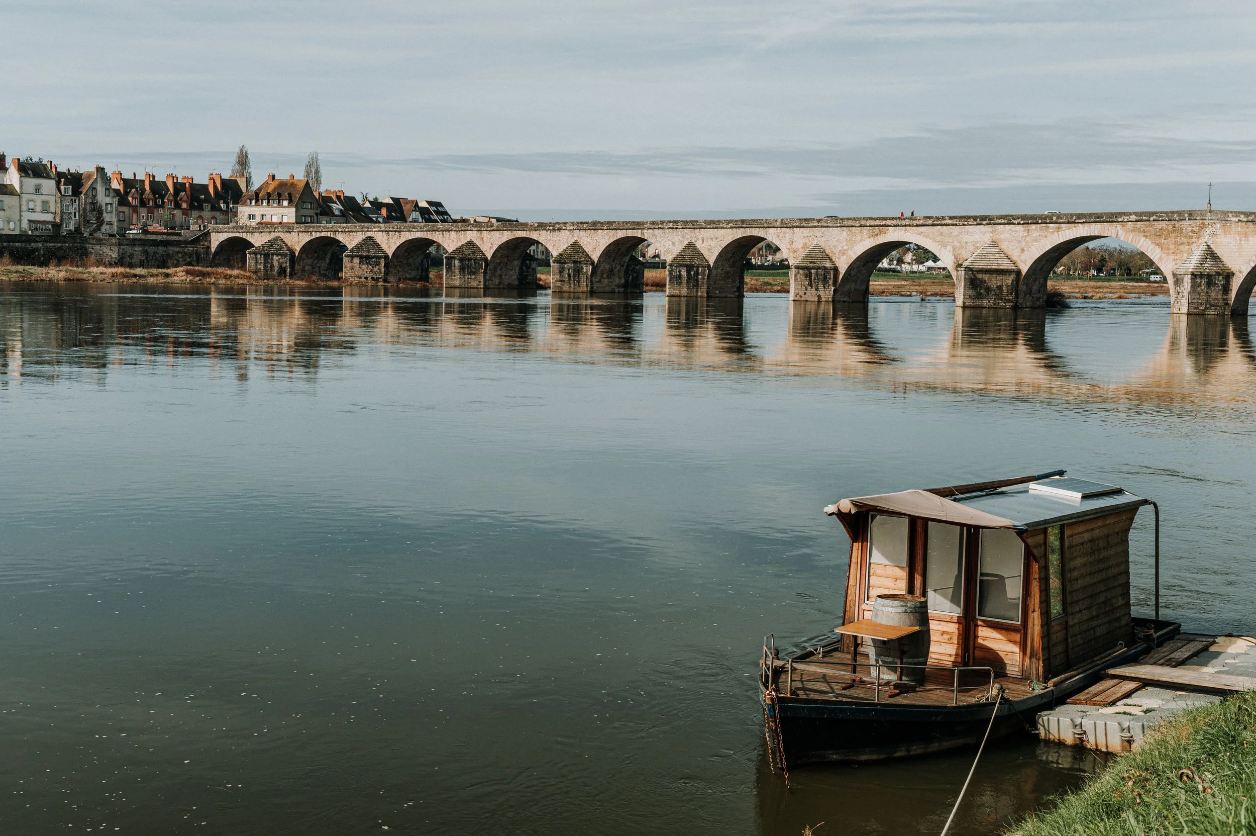 Un bateau en bois amarré au bord d'une rivière avec un pont ancien en pierre en arrière-plan, et des maisons alignées le long du rivage.
