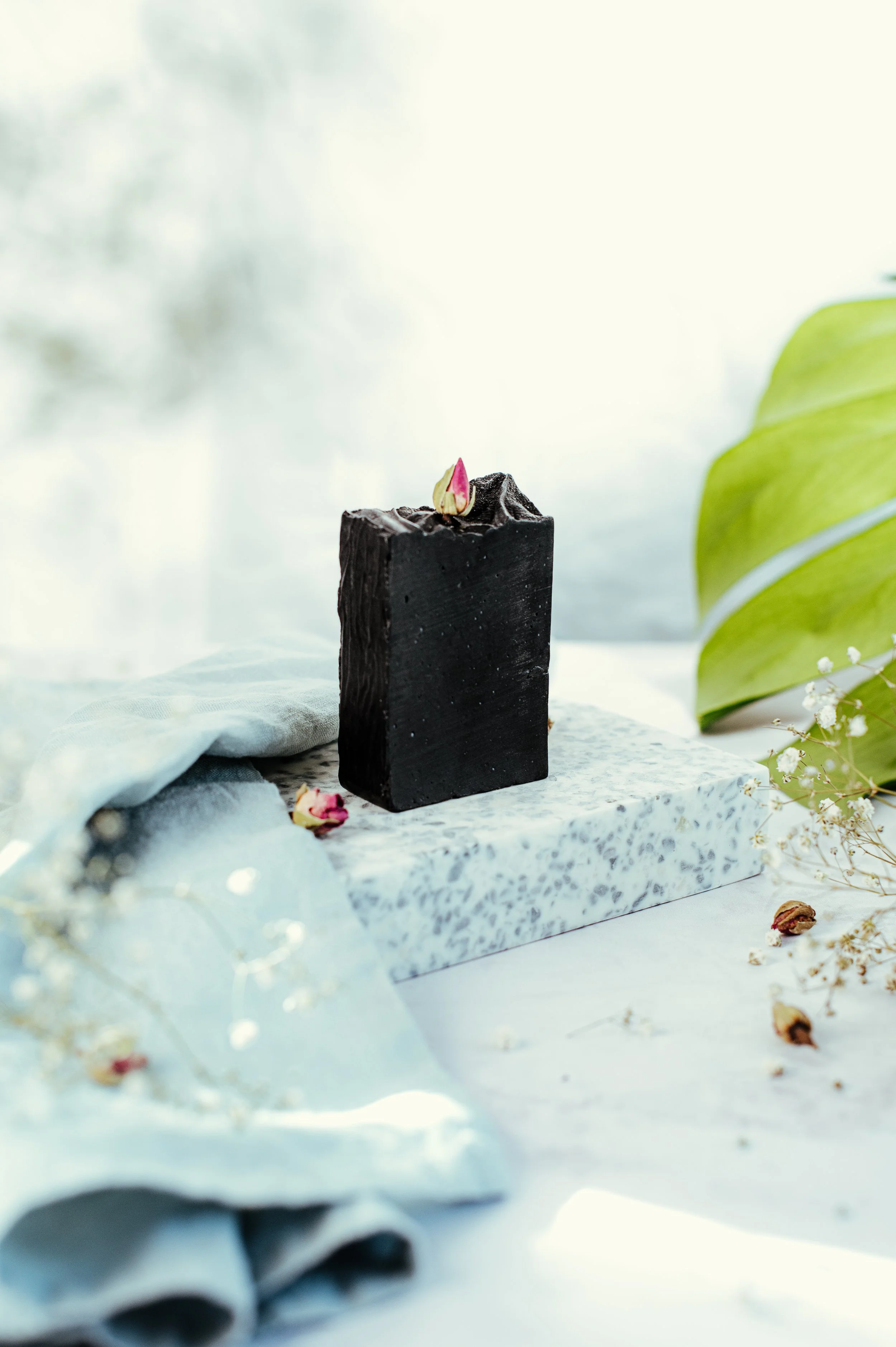 Un gâteau noir rectangular décoré d'une petite feuille de rose, placé sur une plaque de marbre, avec des fleurs séchées et un grand feuillage vert en décor autour.