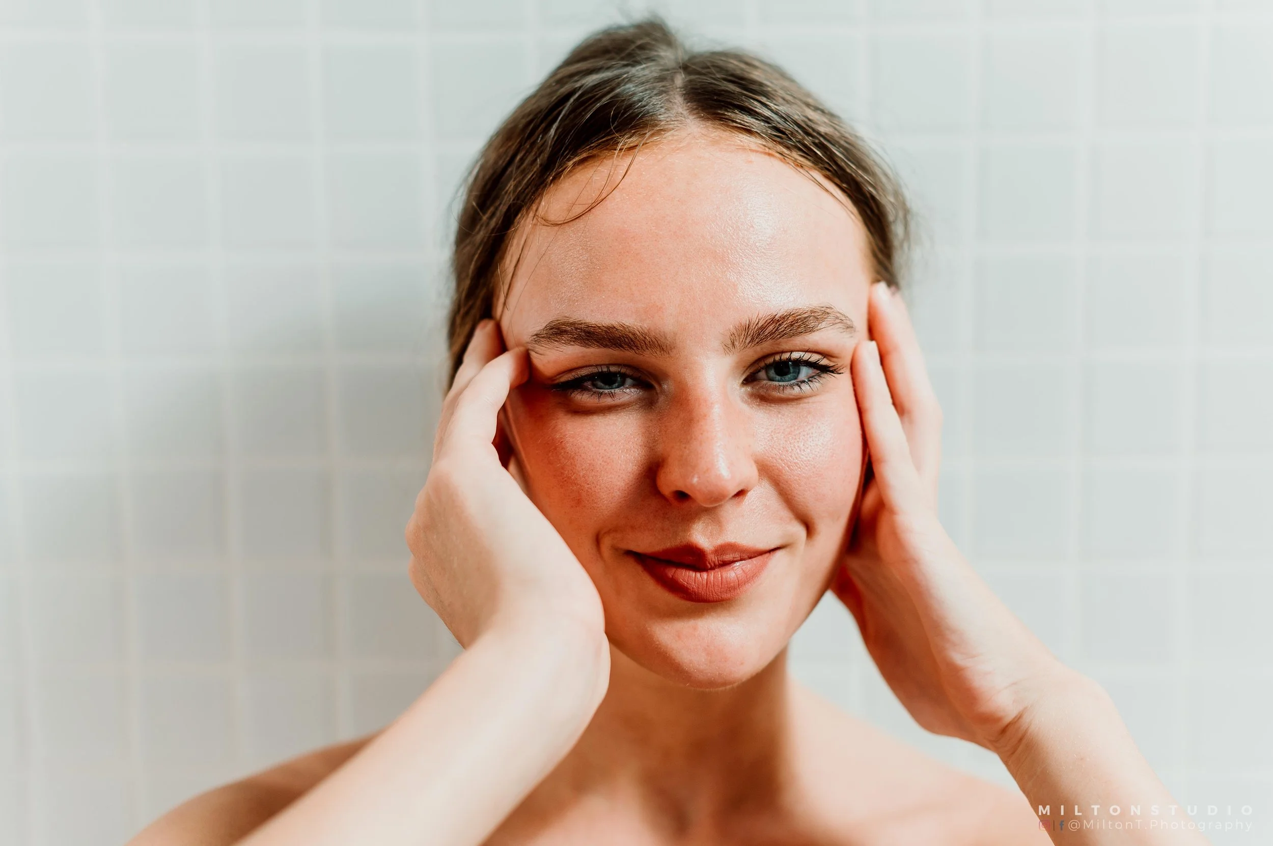 Jeune femme avec un maquillage naturel, souriant et touchant ses tempes, contre un mur en carreaux blancs.