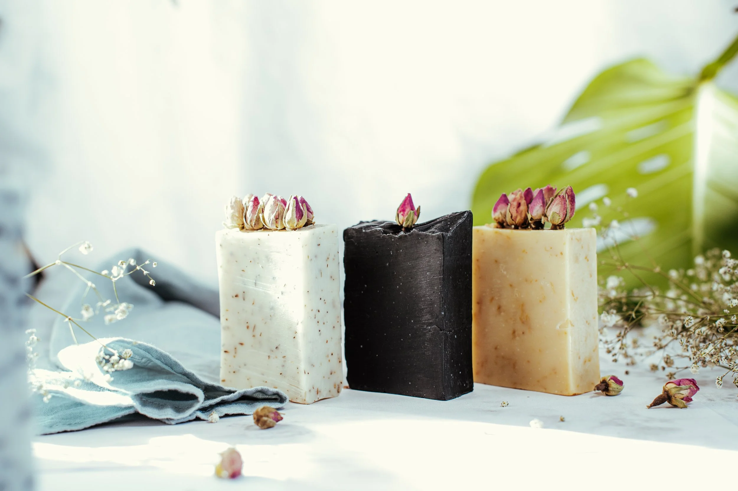 Trois barres de savon posées sur une surface blanche, décorées avec de petites fleurs séchées rose et entourées de fleurs et de feuilles vertes.