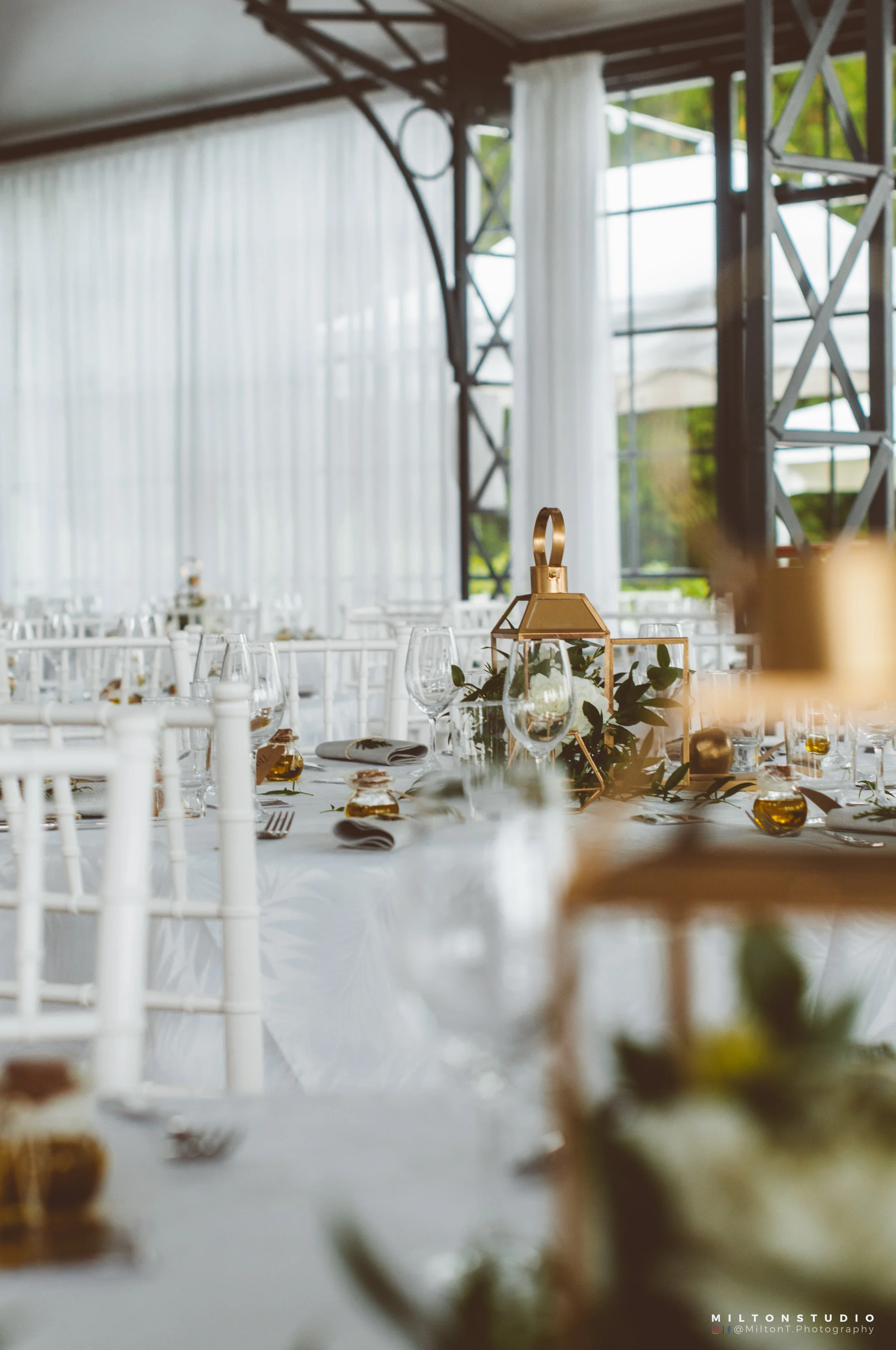 Table de mariage élégante avec décorations en or, bougeoirs, verres à vin, et centres de table avec lanternes et feuillages, dans une salle lumineuse avec des rideaux blancs.