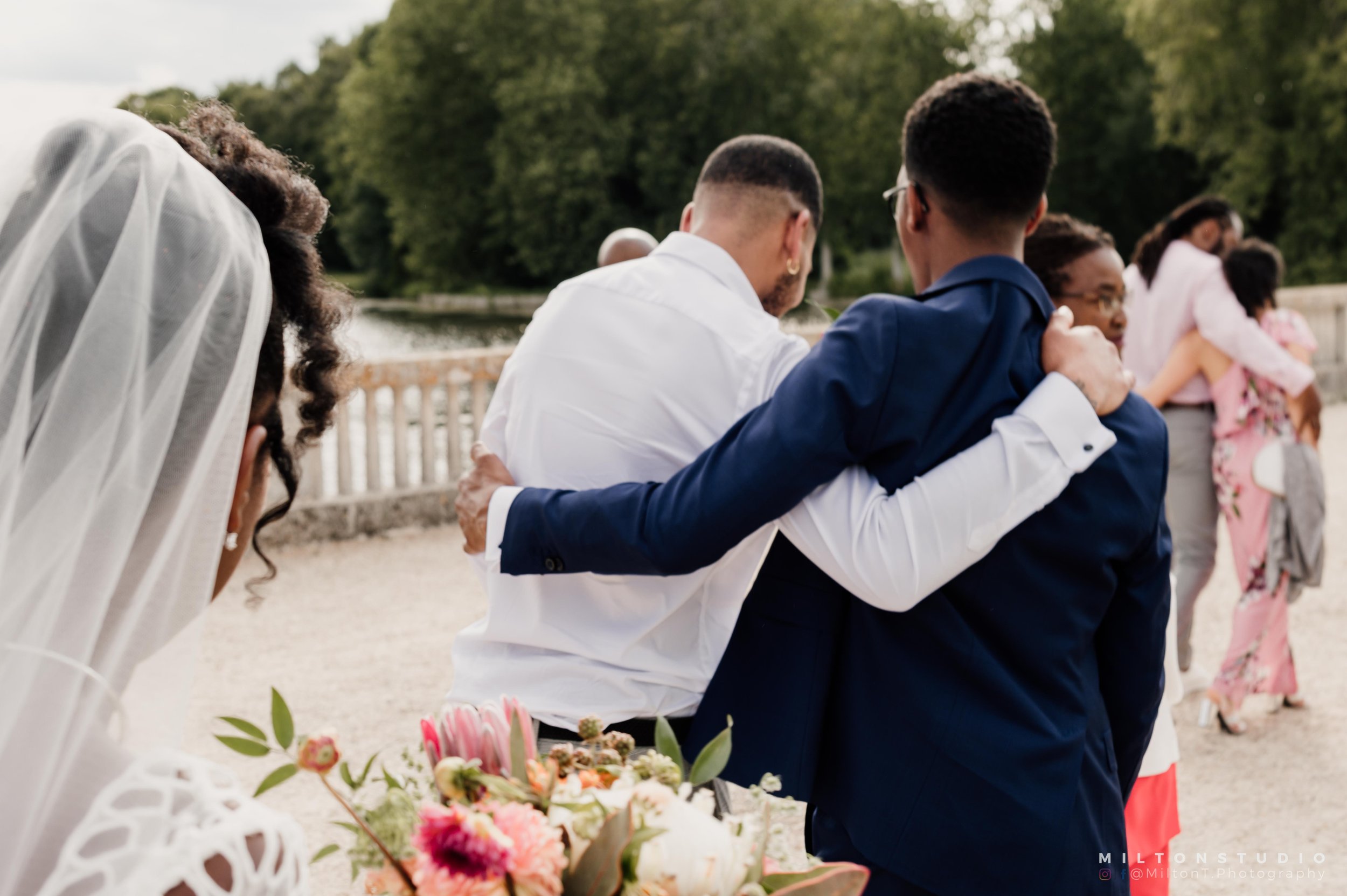Deux hommes en costume se prennent dans les bras lors d'un mariage en extérieur, avec une femme en voile regardant, et d'autres personnes en arrière-plan près d'une rivière et d'arbres.