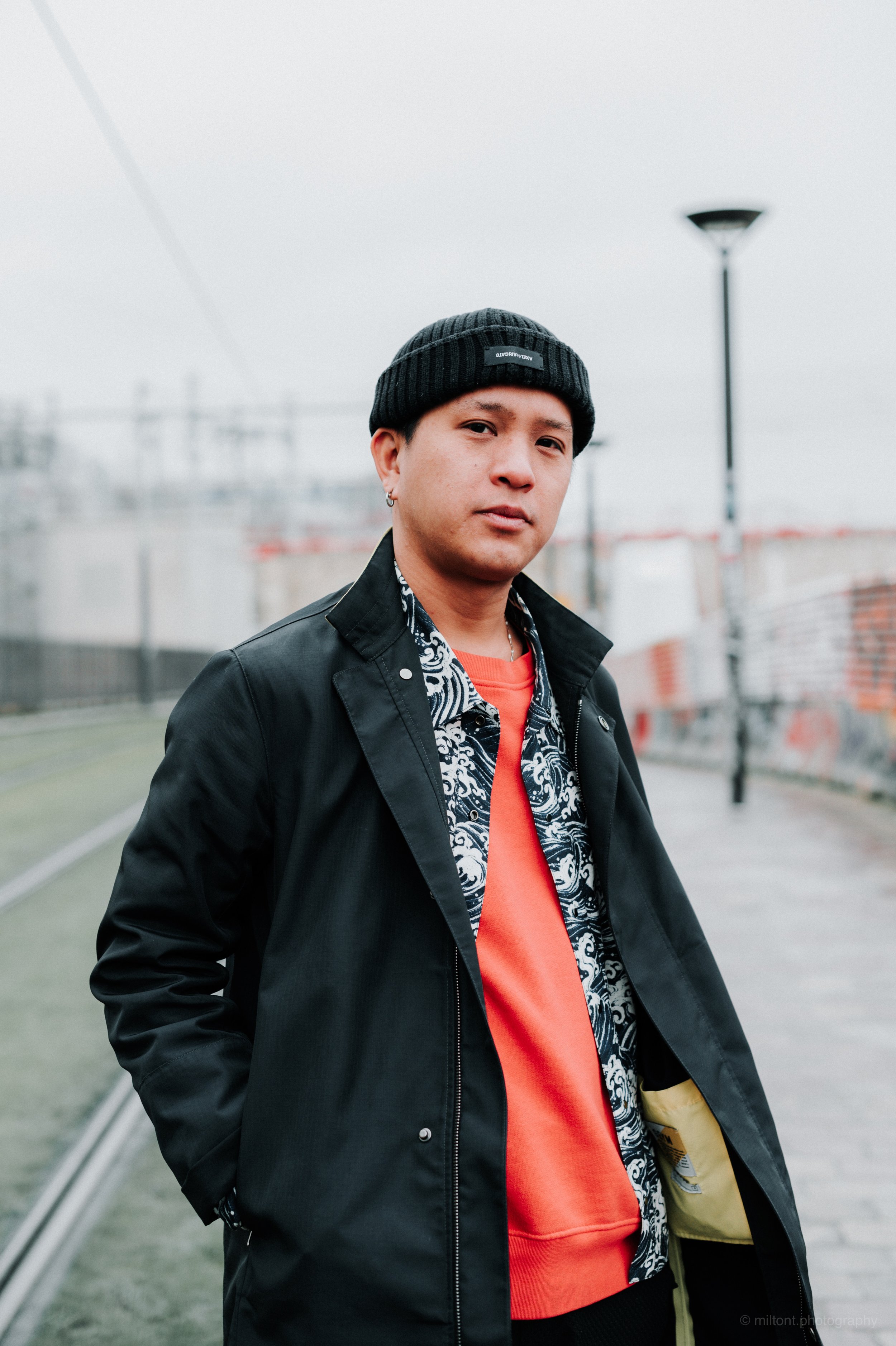 Jeune homme portant un bonnet noir, veste noire, chemise à motifs et sweat orange, en extérieur, à côté d'une voie ferrée, sous un ciel nuageux.