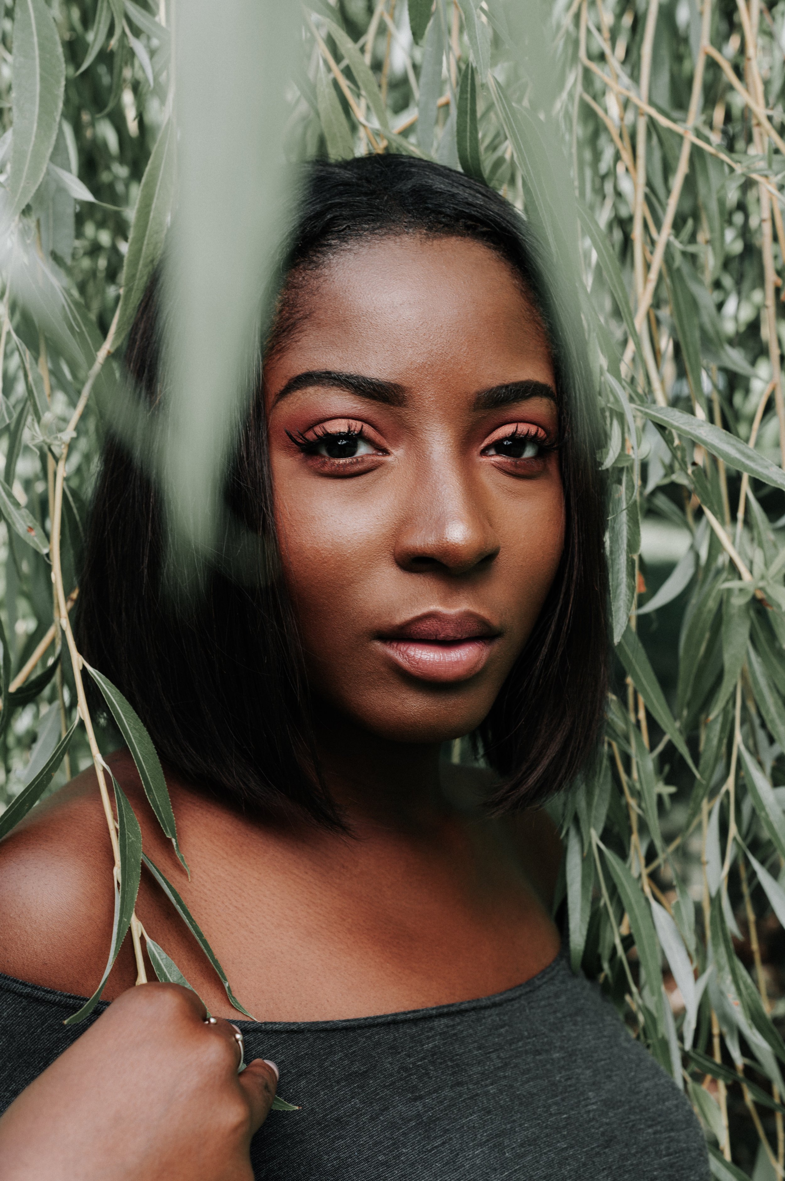 Une femme noire aux cheveux noirs courts, portant un maquillage rose, se tient parmi des feuilles vertes.