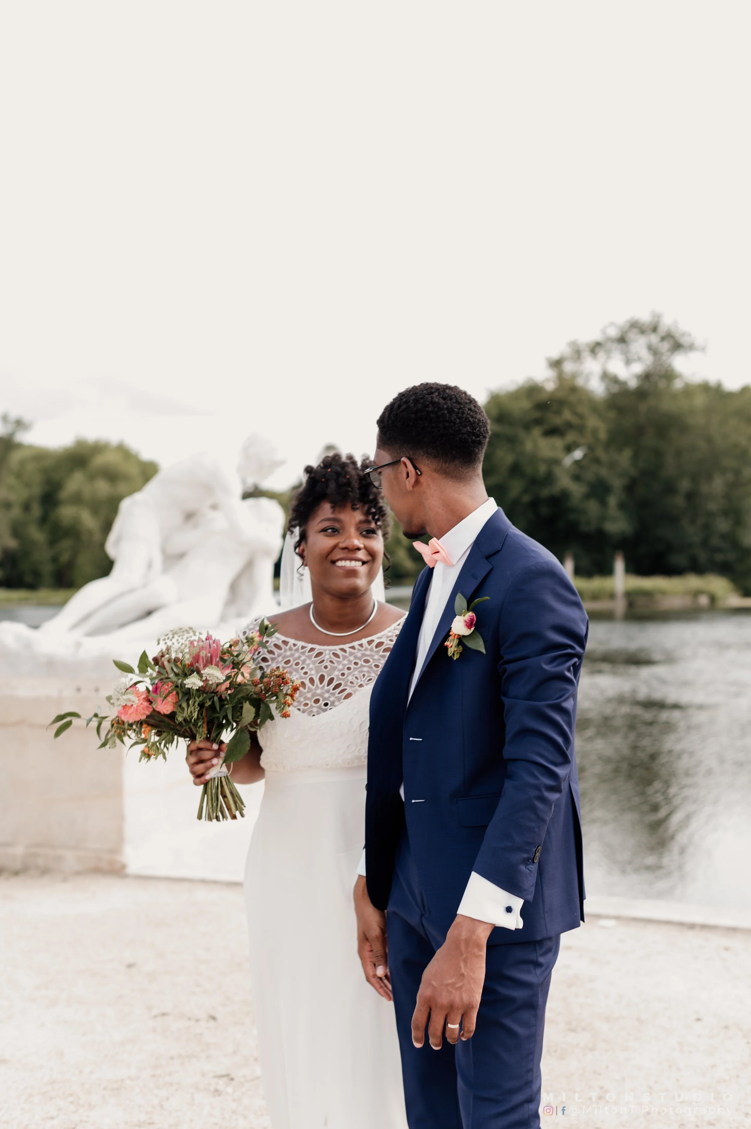 Un couple de mariés devant une fontaine, échangeant un regard et un sourire lors de leur mariage, avec la femme tenant un bouquet de fleurs.