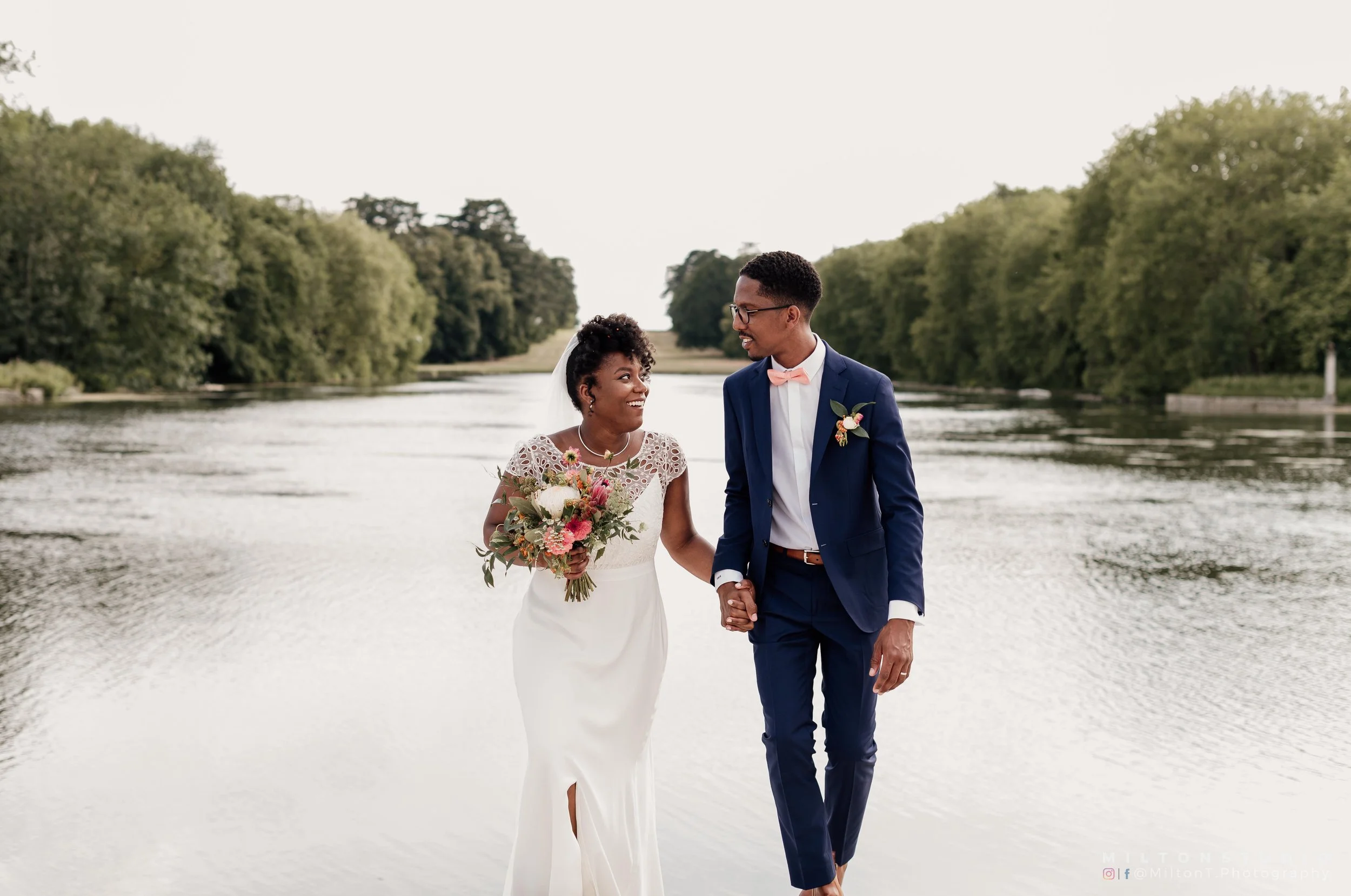 Un couple de mariés qui marche sur une rivière, tenant la main, avec un paysage verdoyant en arrière-plan.