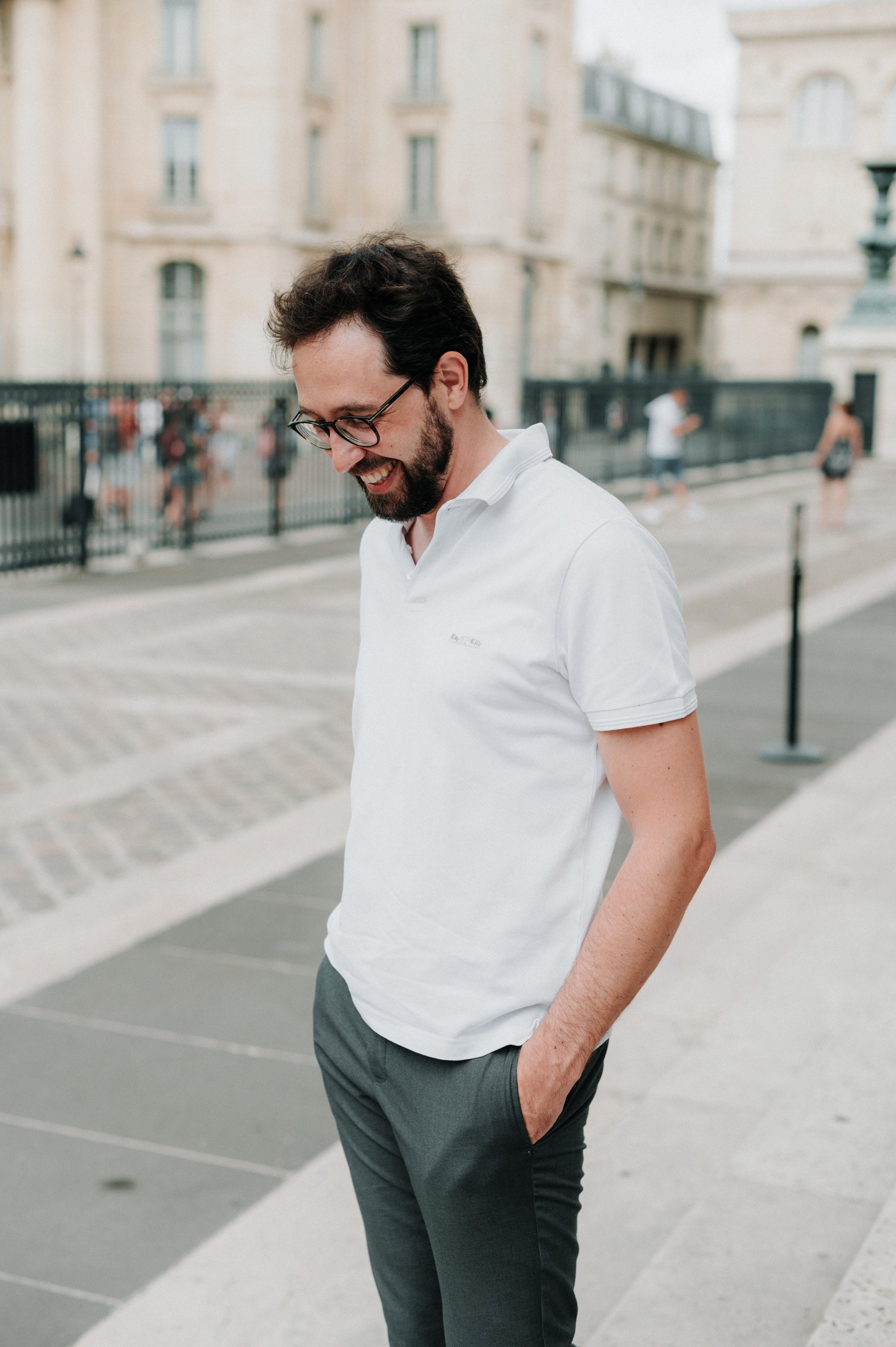 Un homme avec des lunettes, une barbe et une chemise blanche sourit et regarde vers le bas dans une rue pavée en arrière-plan d'un bâtiment en pierre
