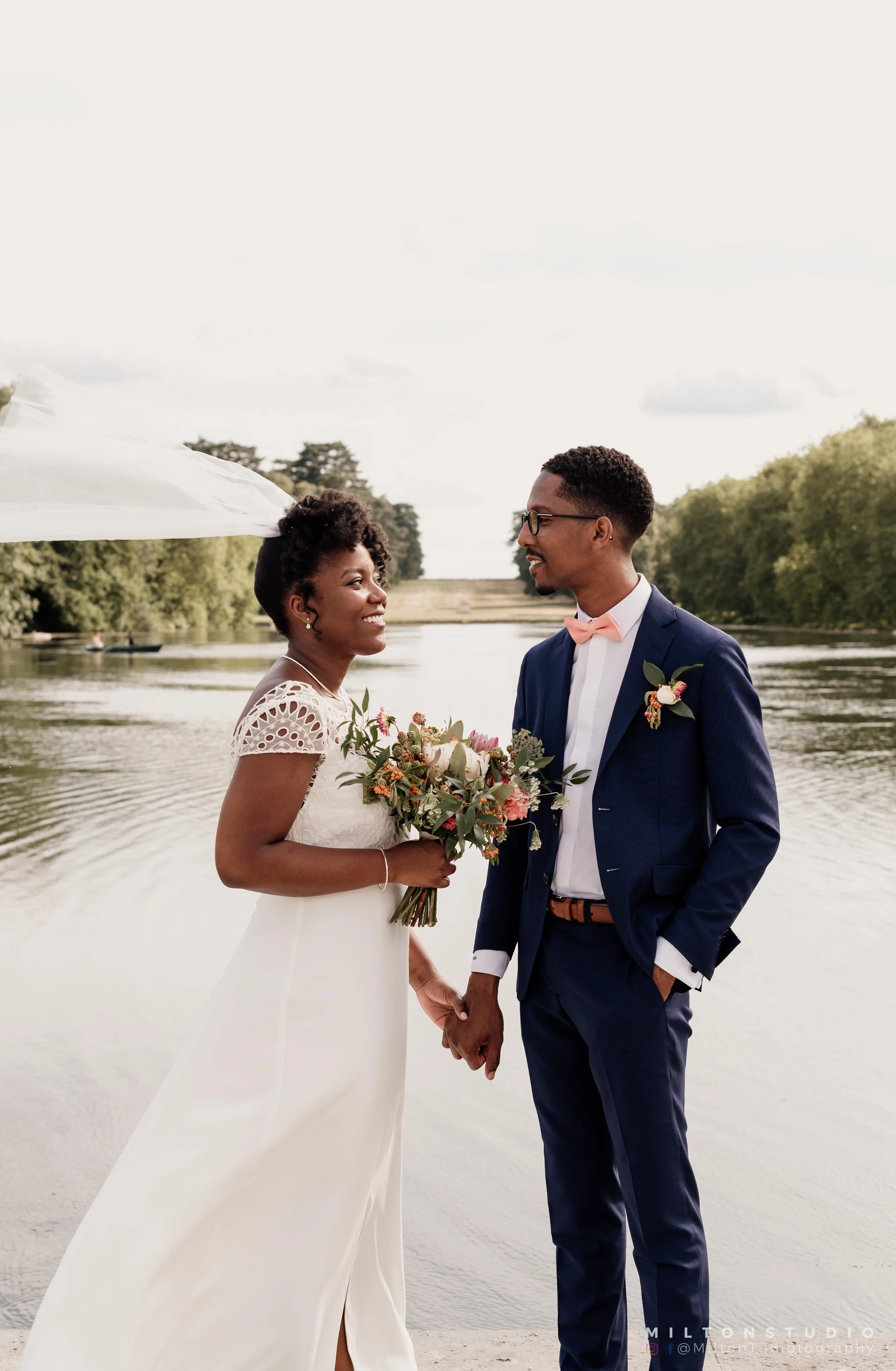 Un couple de mariage, la femme en robe blanche et le homme en costume bleu foncé, se tenant par la main près d'une rivière, avec un bouquet de fleurs dans les mains de la femme, un ciel partiellement nuageux en arrière-plan.