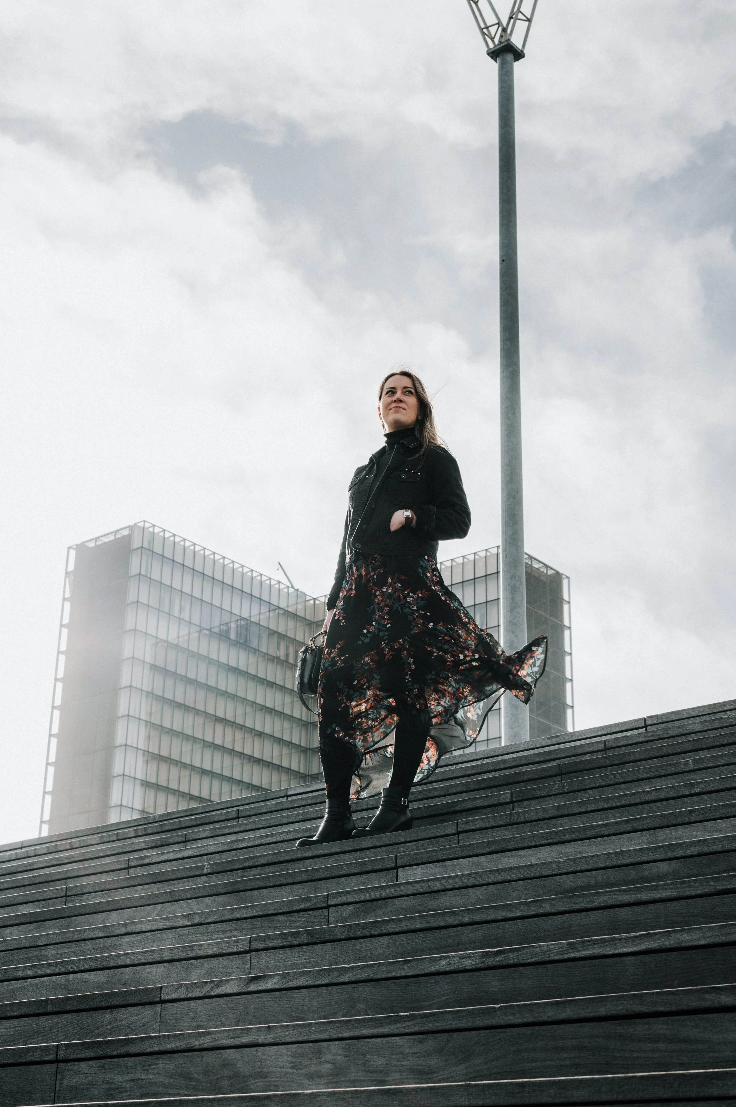 Femme debout sur des escaliers en bois, portant une robe longue à motifs floraux, une veste noire et des bottes, sous un ciel nuageux, avec des bâtiments modernes en arrière-plan.