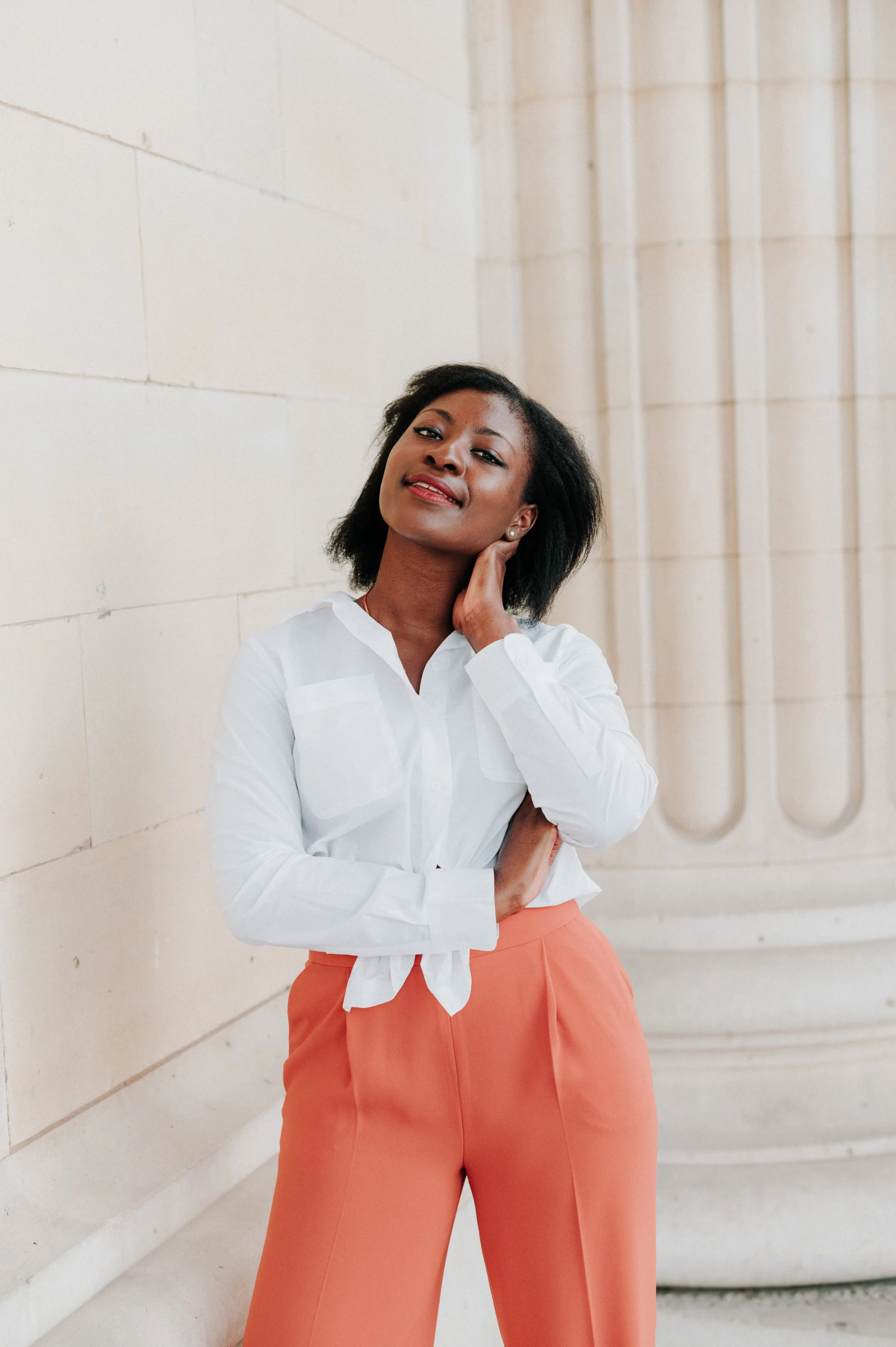 Une femme noire en chemise blanche et pantalon orange clair, posant contre un mur en pierre beige avec une colonne ionique visible à l'arrière.