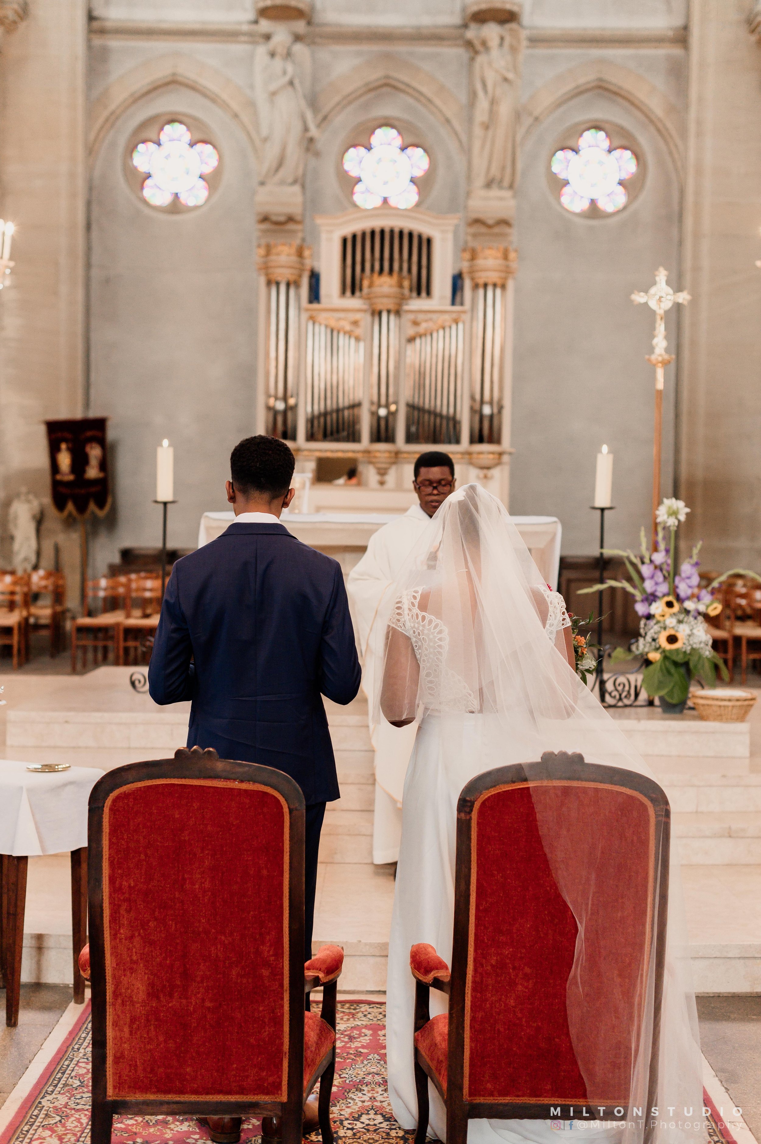 Un couple en mariage, vu de derrière, au nœud papillon et d'une robe blanche avec un voile, pendant une cérémonie religieuse dans une église architecturale.