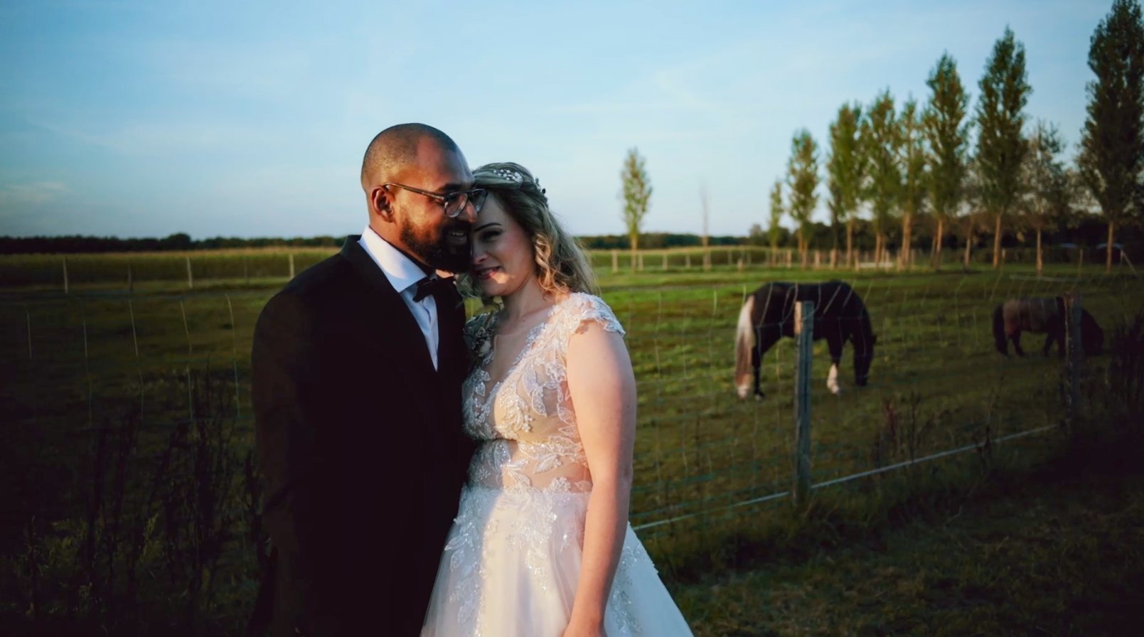 Un couple de mariés dans une ferme, avec des chevaux en arrière-plan, lors du coucher de soleil.