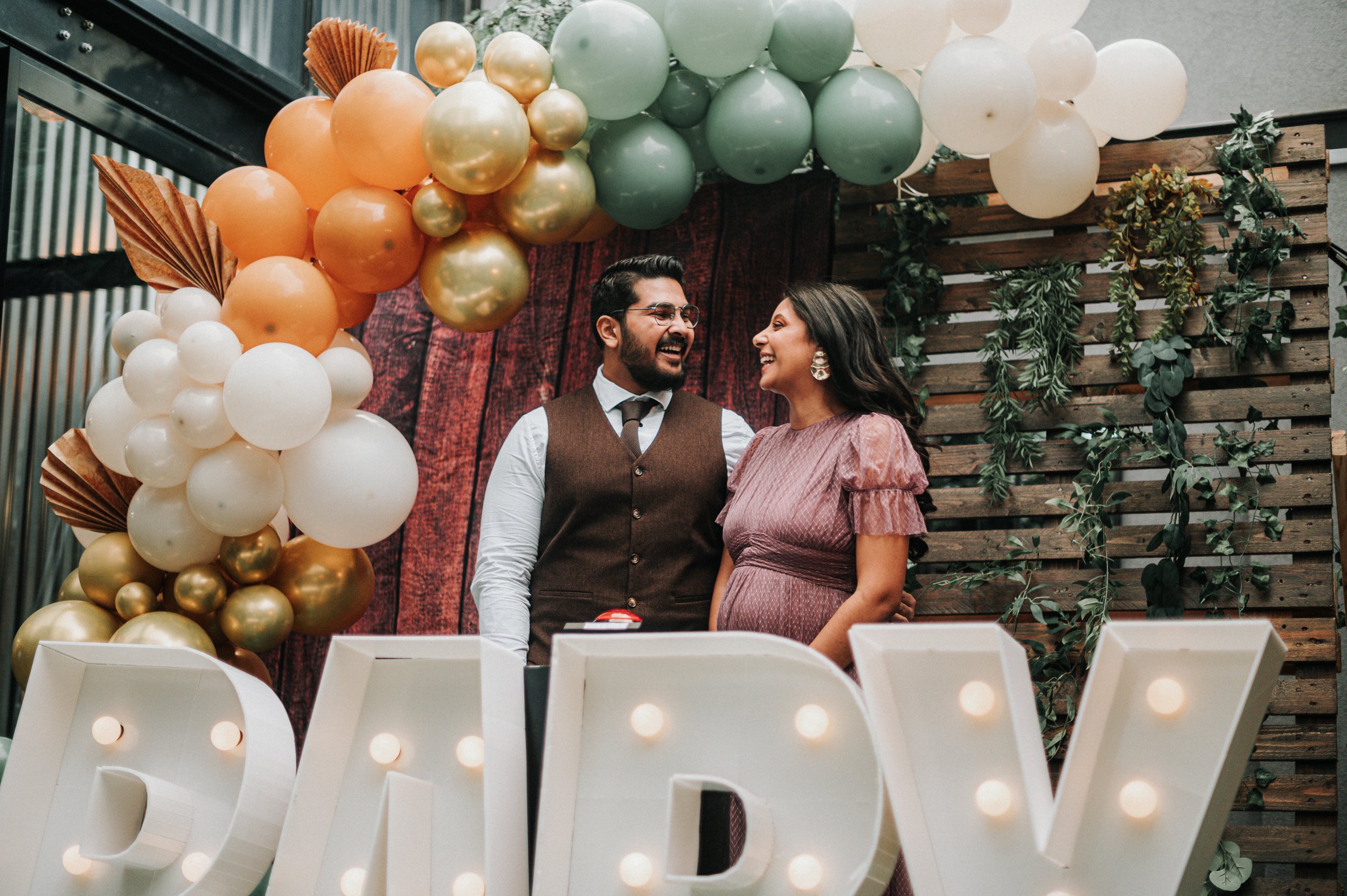 Un couple souriant lors d'une fête ou d'une célébration, entouré de décorations de ballons et de lettres lumineuses « M» et « D ». La femme porte une robe mauve et des boucles d'oreilles, l'homme porte une chemise blanche, un gilet marron et une crav