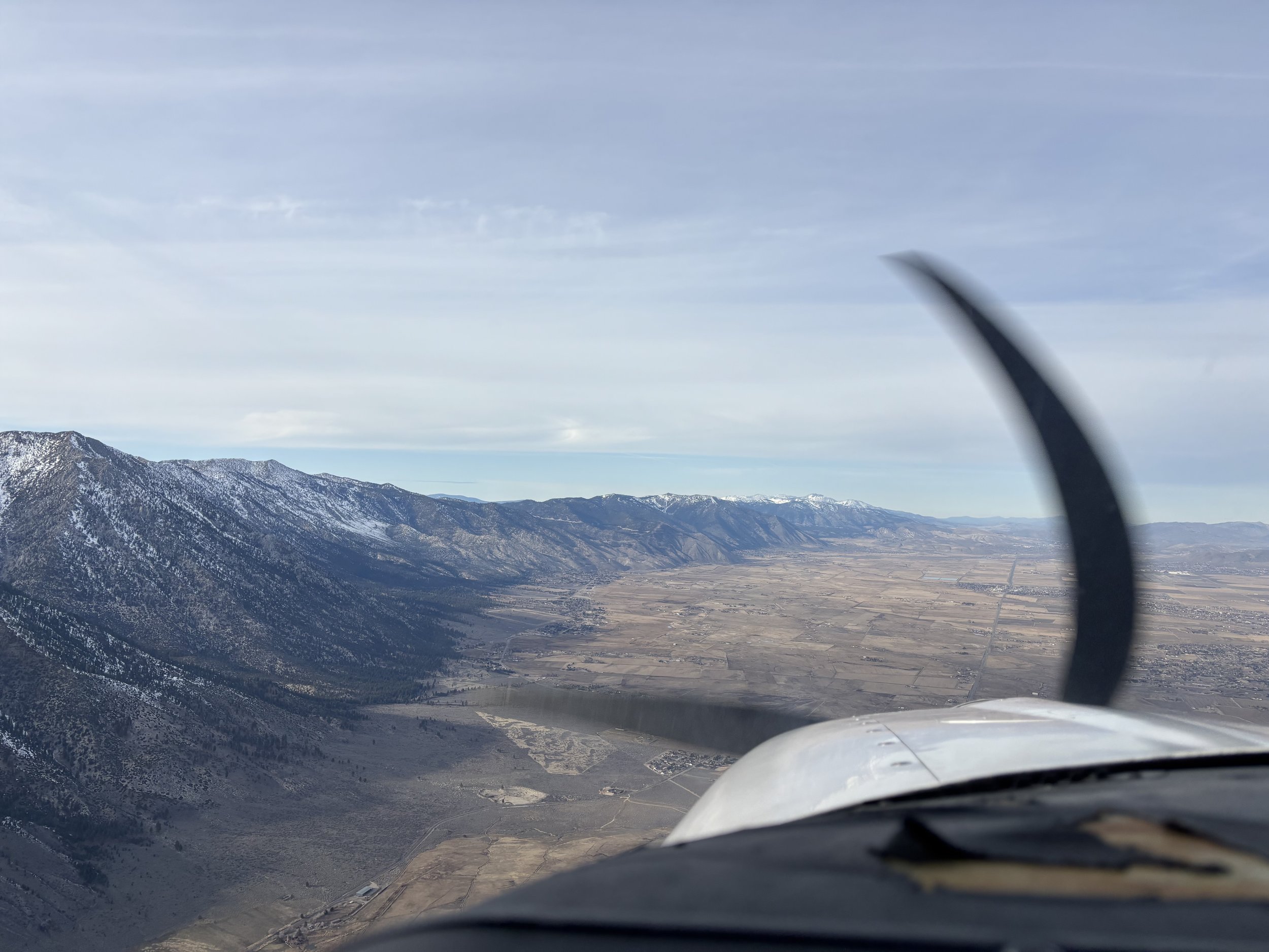 View from an aircraft showing a mountain range with snow patches, open land, and distant snow-capped peaks under a partly cloudy sky.