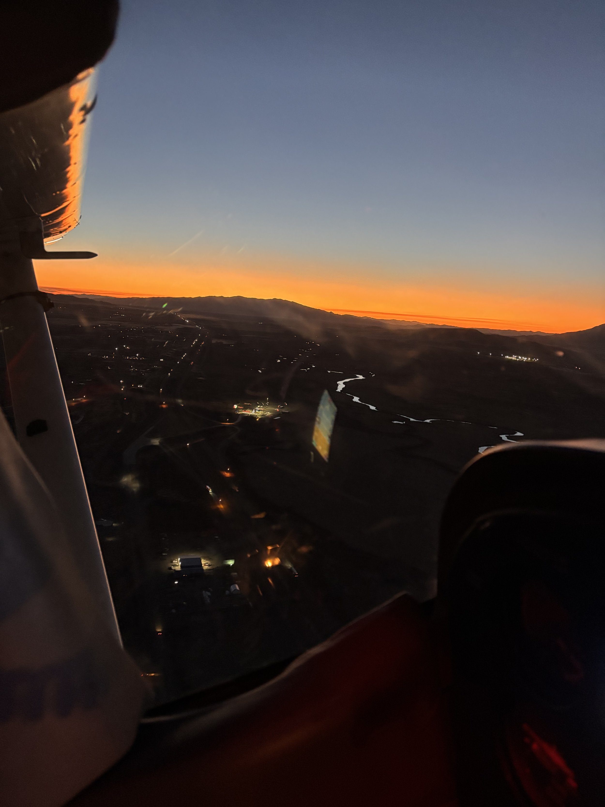 View of a sunset from an airplane cockpit, showing the sky, mountains, and a river below.