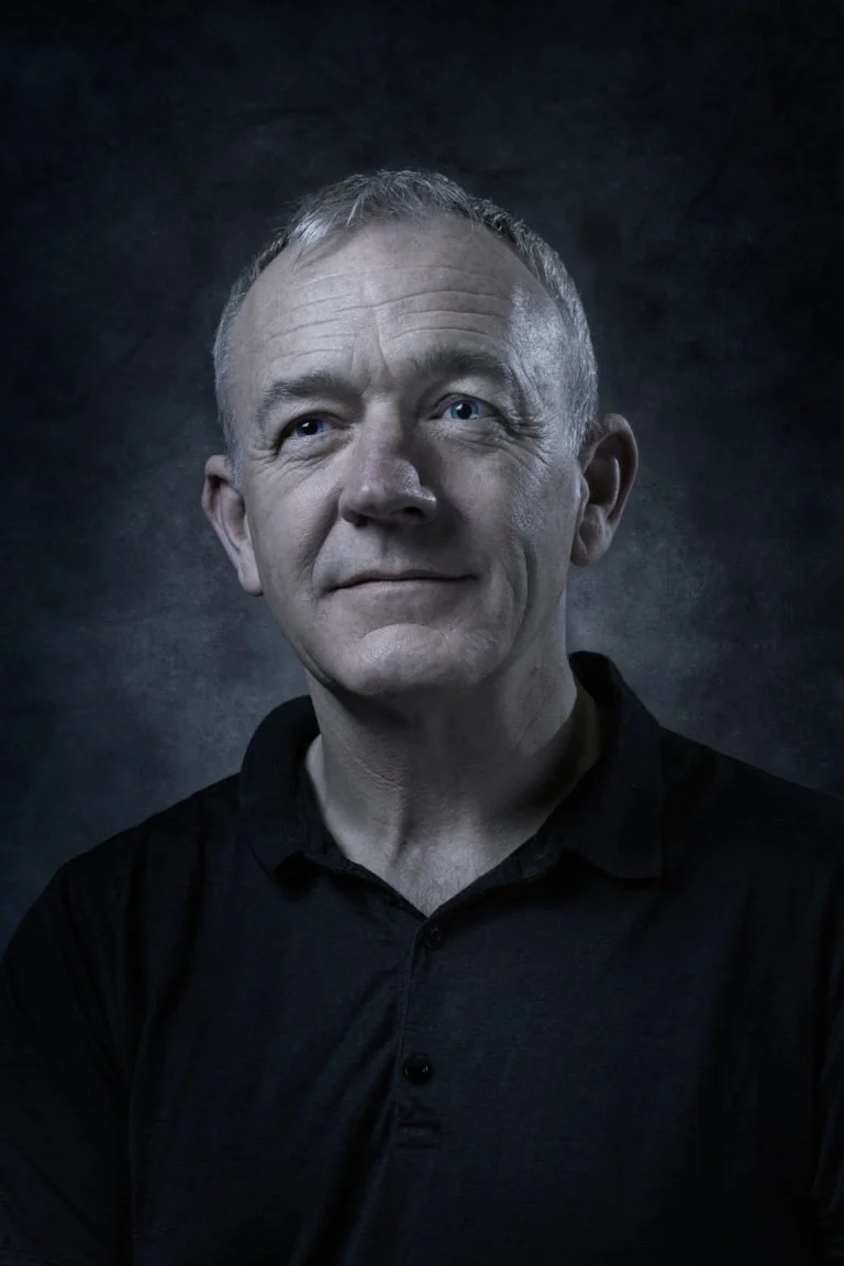 A man with a beard and short hair standing outdoors in a wooded area, holding a camera with a large telephoto lens.