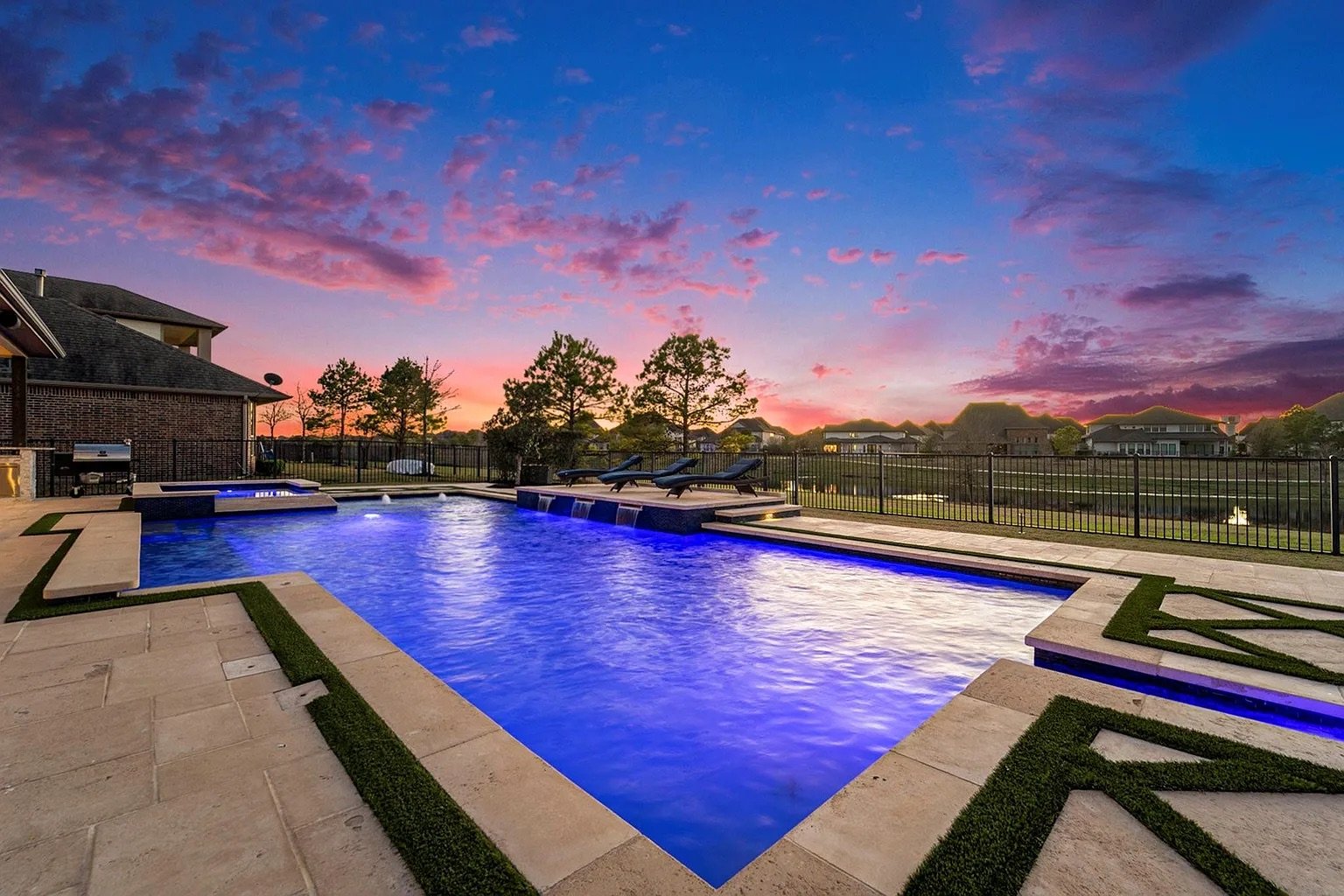 An illuminated backyard swimming pool at sunset with a landscape of houses and trees in the background.