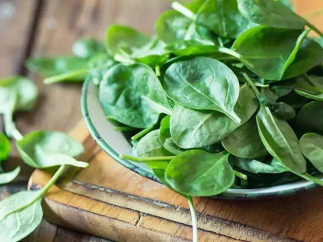 Fresh spinach leaves in a bowl with some leaves scattered on a wooden surface.