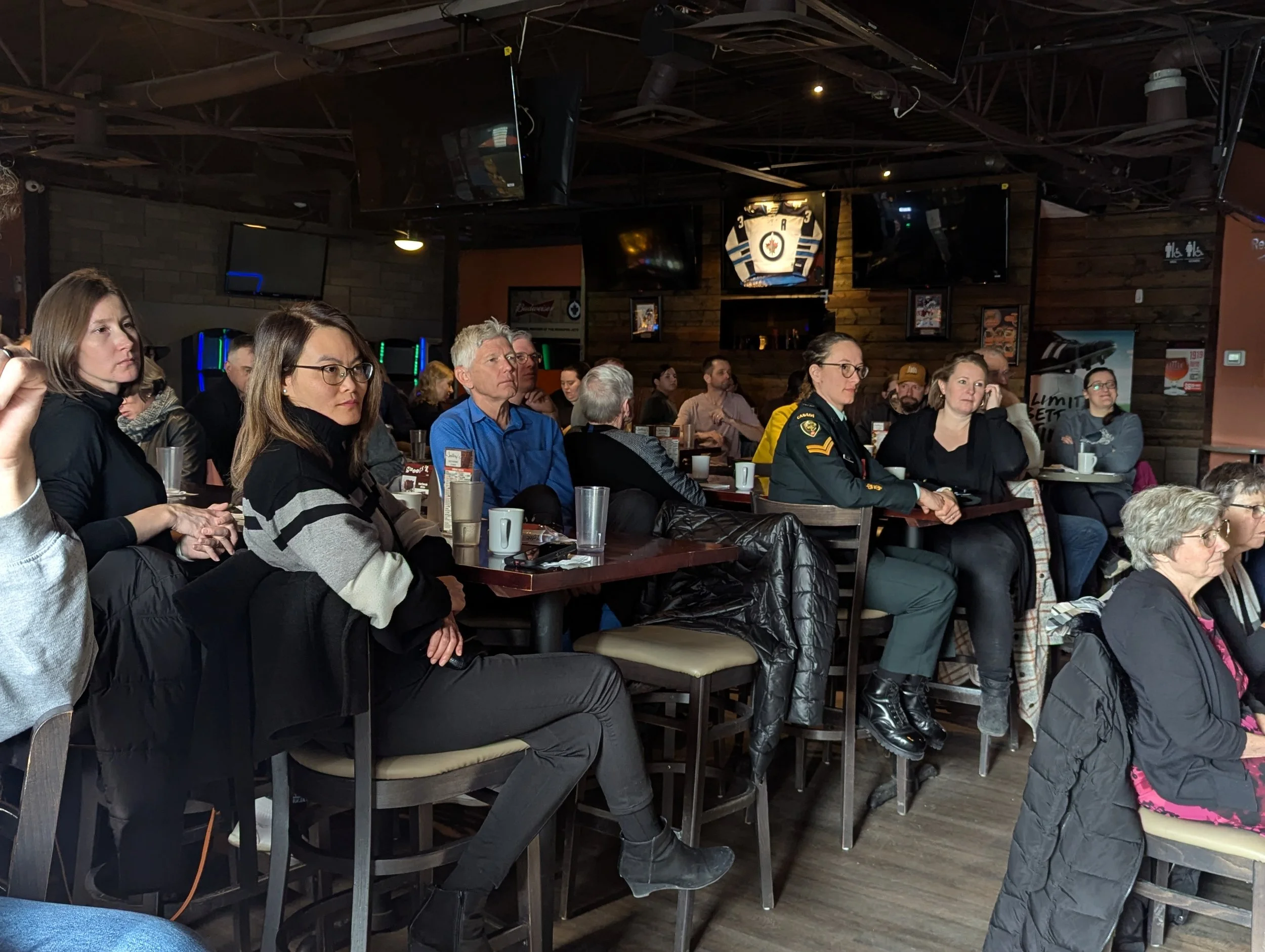 Group of people seated at tables in a dimly lit sports bar, watching a presentation or event on multiple TV screens.