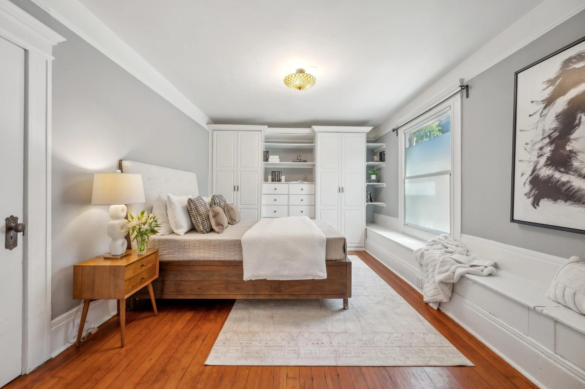 A bedroom with a wooden bed, white bedding, and multiple pillows, a wooden nightstand with a lamp and a small flower vase, white built-in cabinets and shelves, a large window with frosted glass, and a wall art with a horse painting.