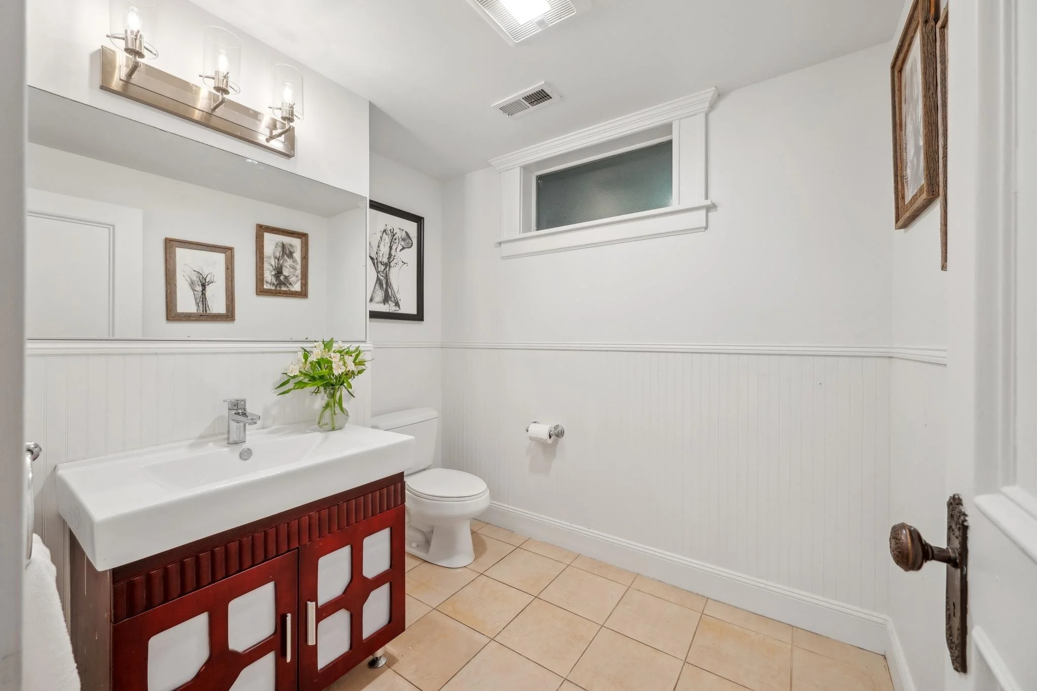 Small bathroom with white walls, a tile floor, a white sink with a wooden cabinet, a vase with flowers, a mirror, framed artwork, a window, a toilet, and a towel holder.
