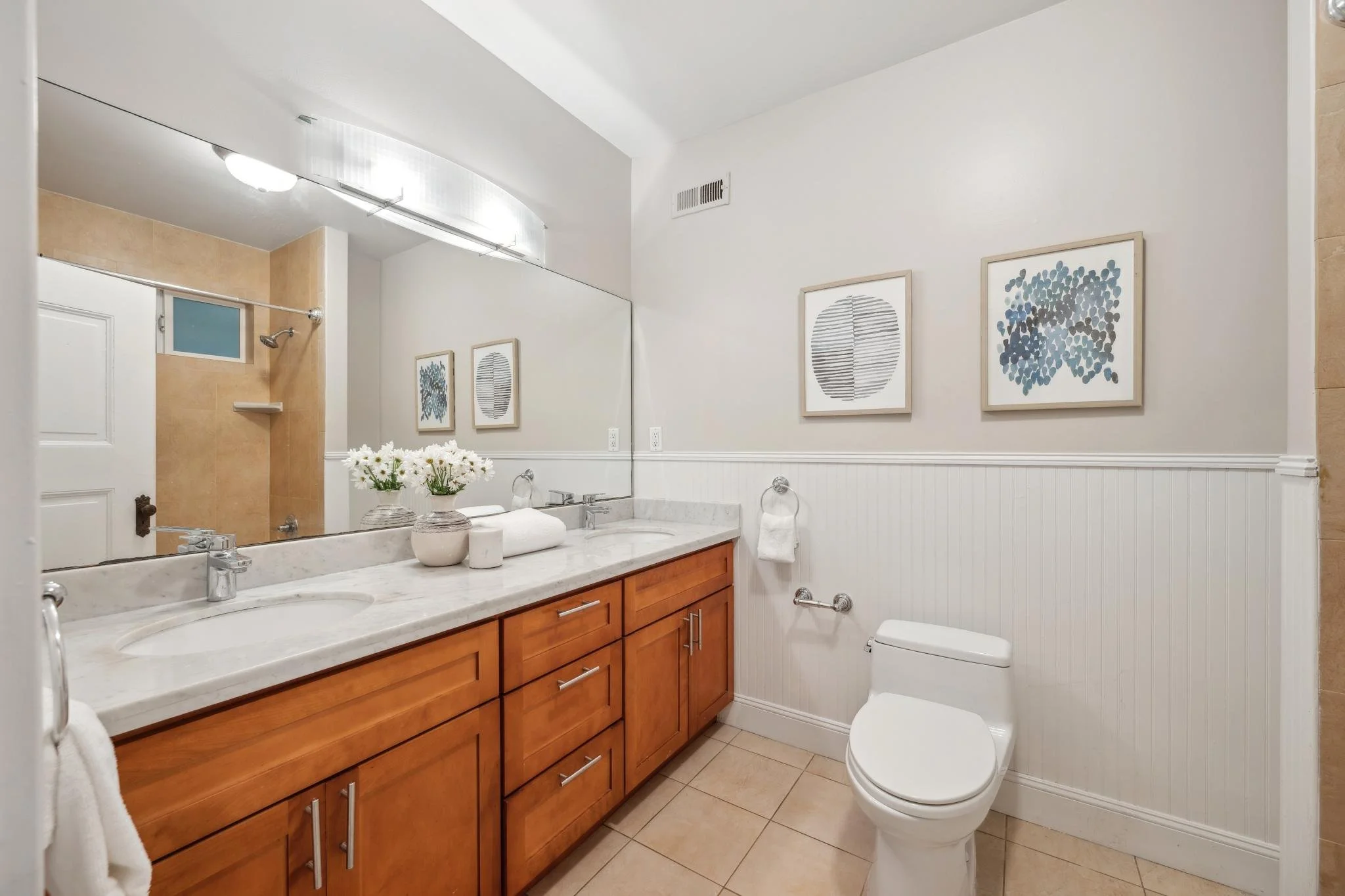 A bathroom featuring a large mirror above a marble countertop with a vase of white flowers and bathroom accessories, wooden cabinets below, a toilet with a towel nearby, and framed modern art on the wall.