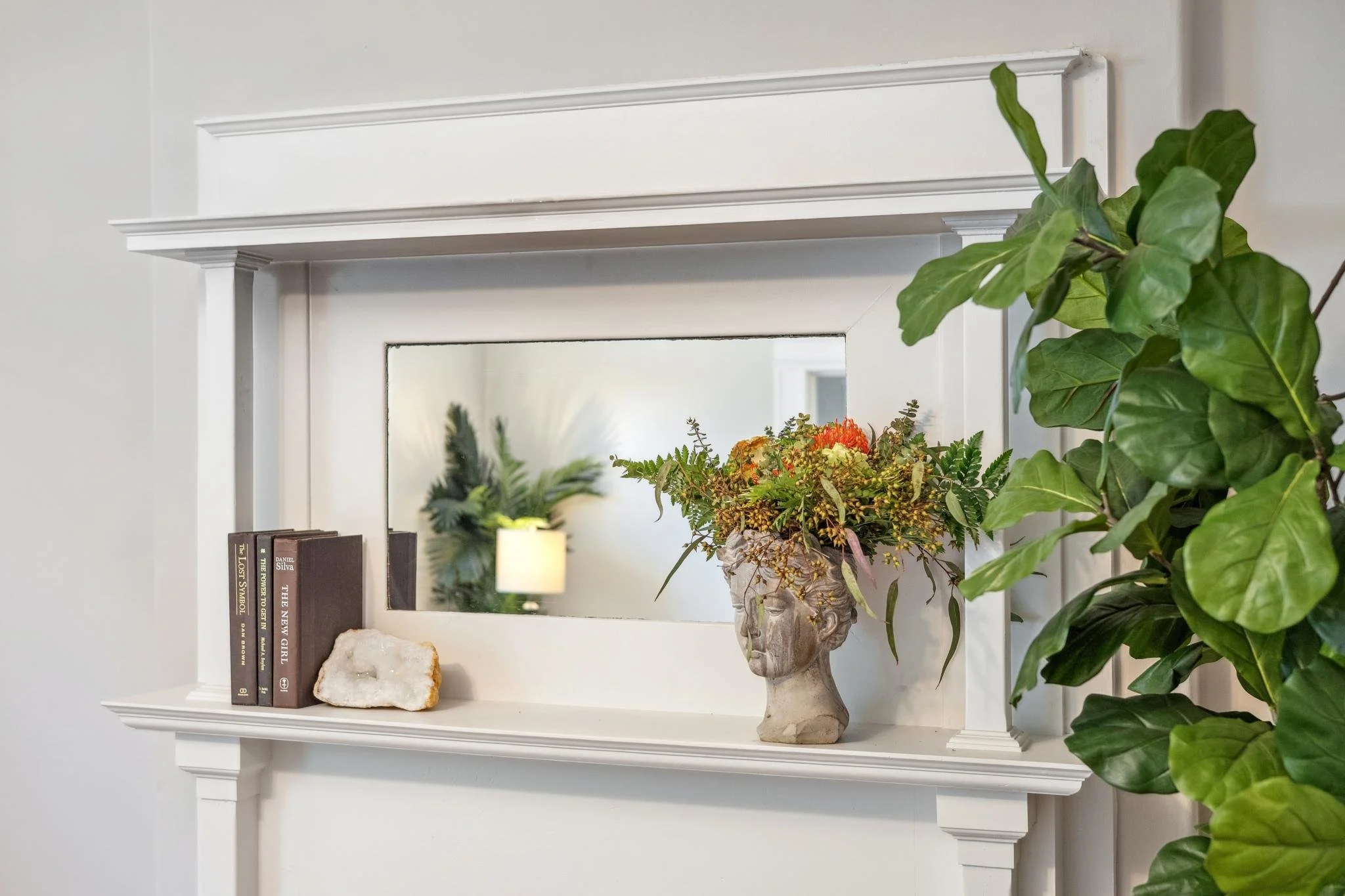 Decorative white shelf with books, a rock, a ceramic face-shaped vase with flowers, and a large houseplant in a white room, with a mirror reflecting a potted plant and a lamp.