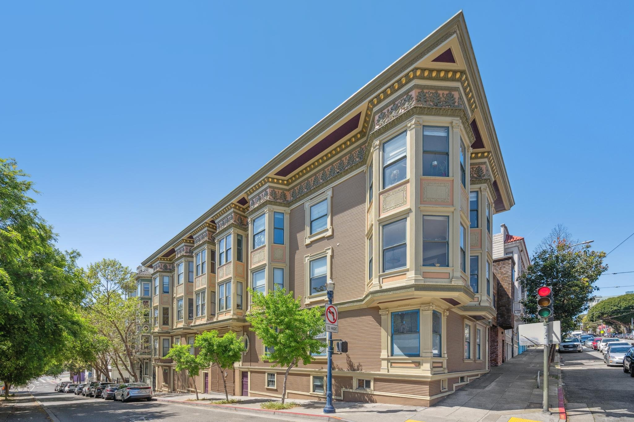 A four-story historic apartment building with ornate decorative trim and bay windows on a sunny city street corner, with parked cars and green trees in the foreground.