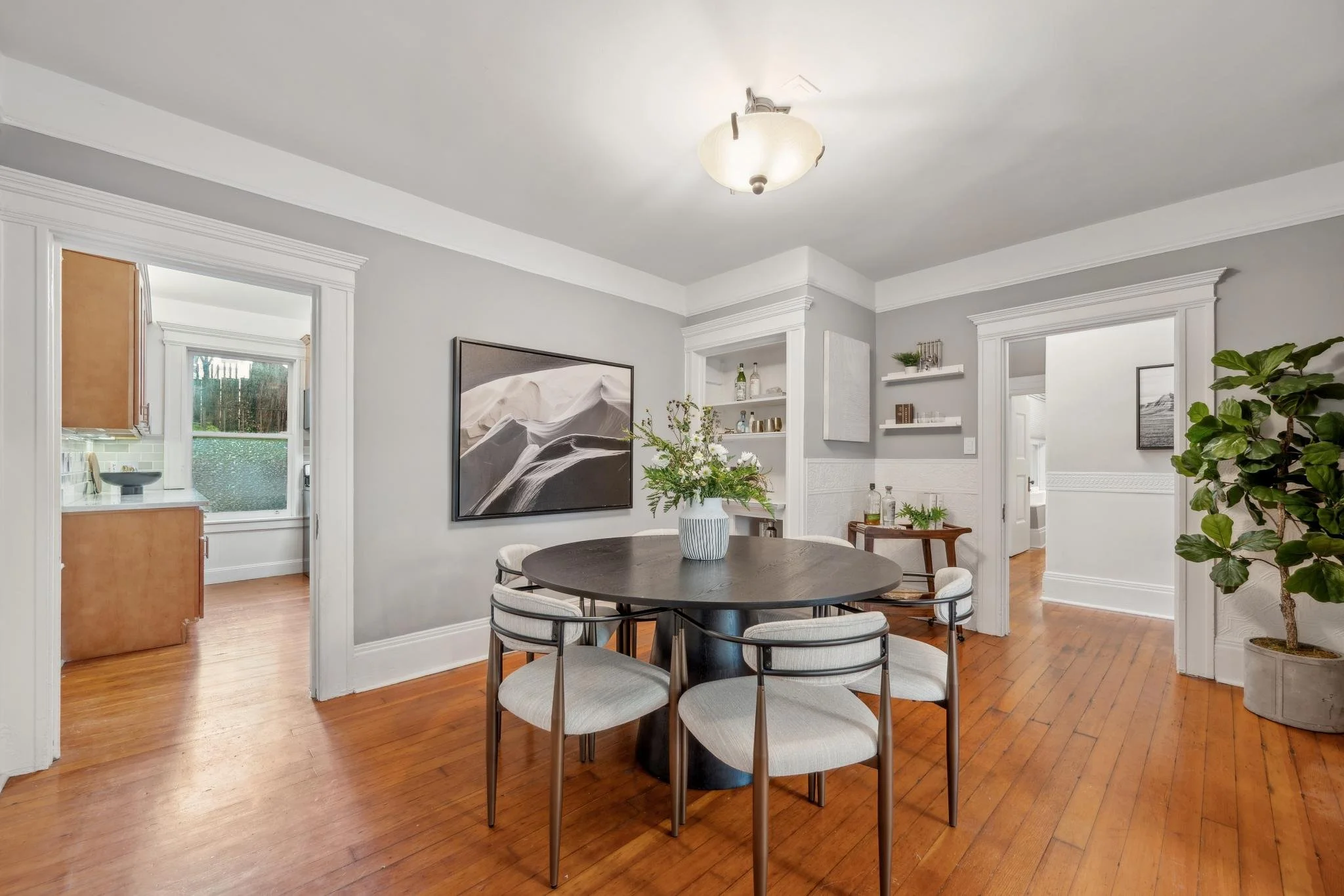 Dining room with round black table, six white upholstered chairs, a white vase with green and white flowers, wooden floors, gray walls, and white trim. Adjoining kitchen with window and cabinet visible.