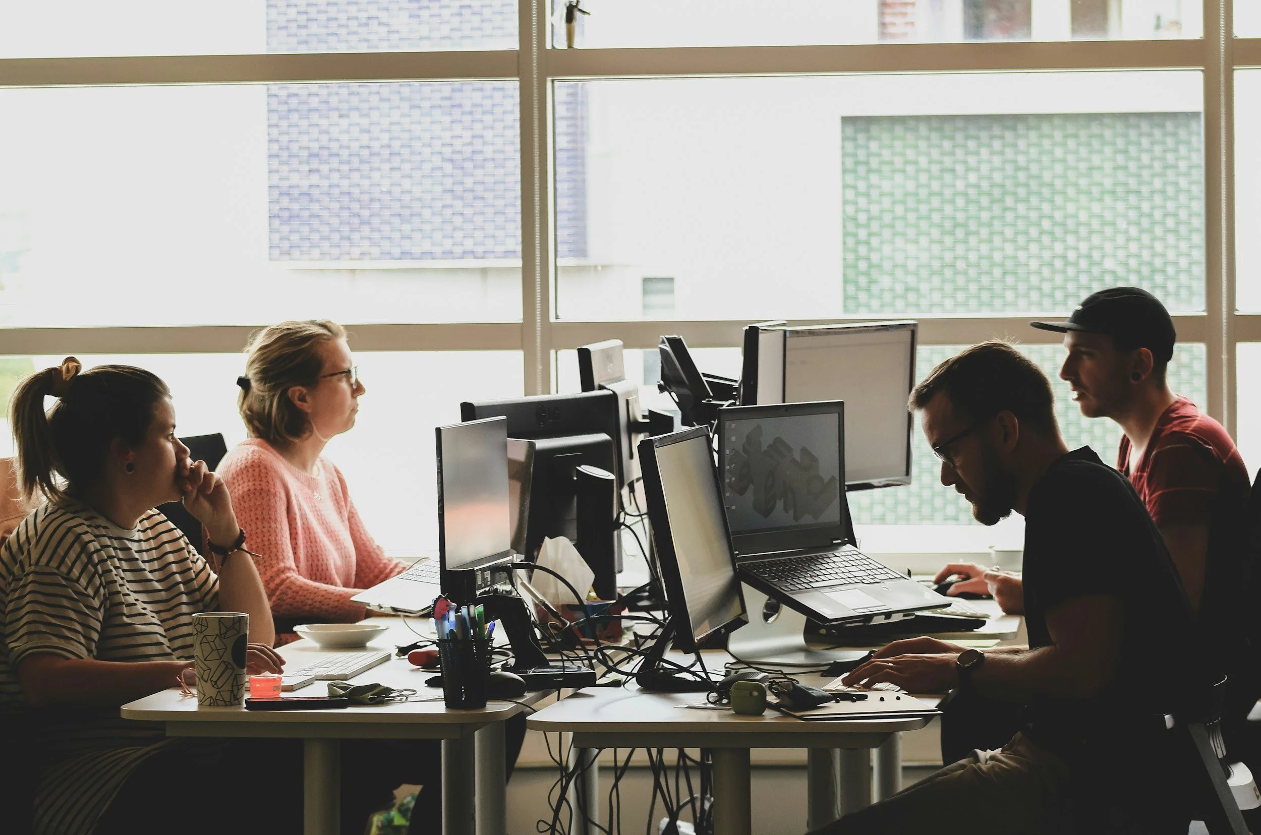 Group of people working at computers in an office environment with natural light through large windows.