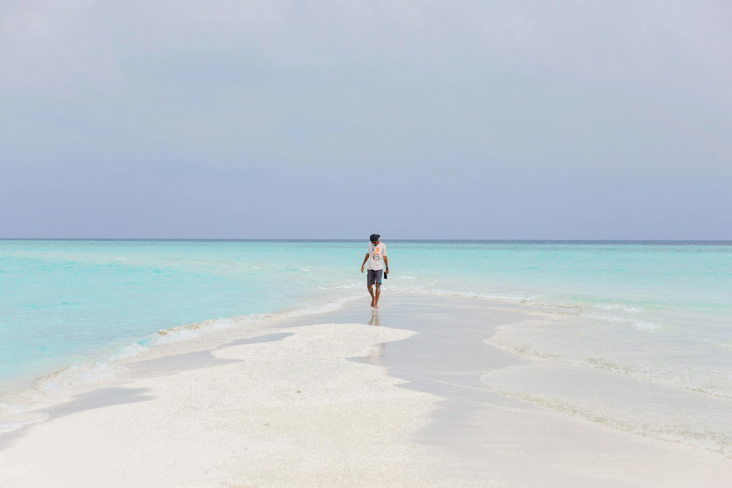A person walking along a narrow strip of sandy beach into turquoise ocean waters under a cloudy sky.
