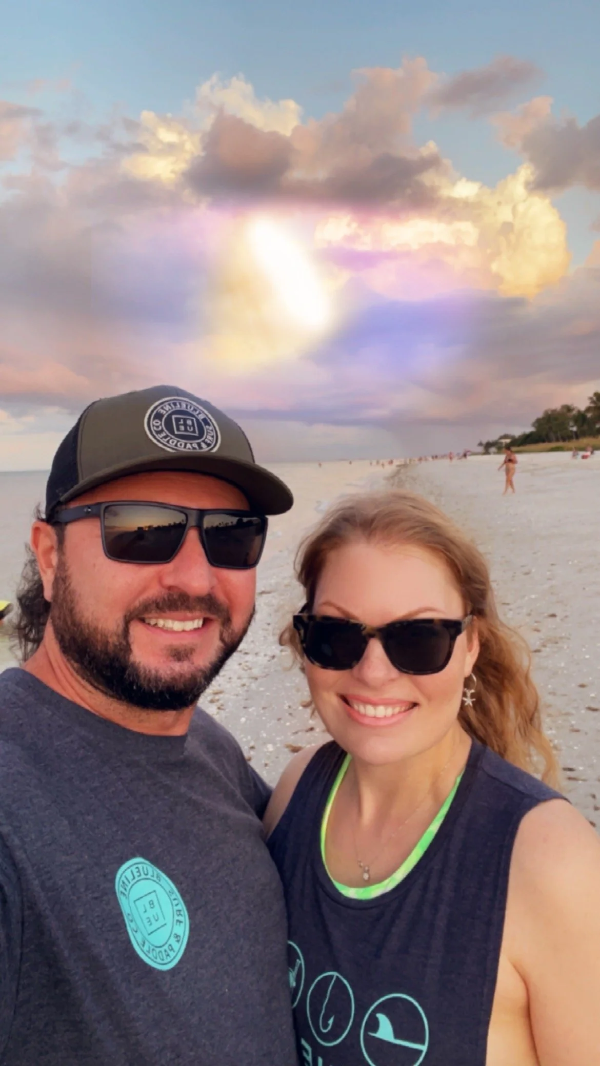 Couple taking a selfie on a beach at sunset, wearing sunglasses, with cloudy sky and people in the background.