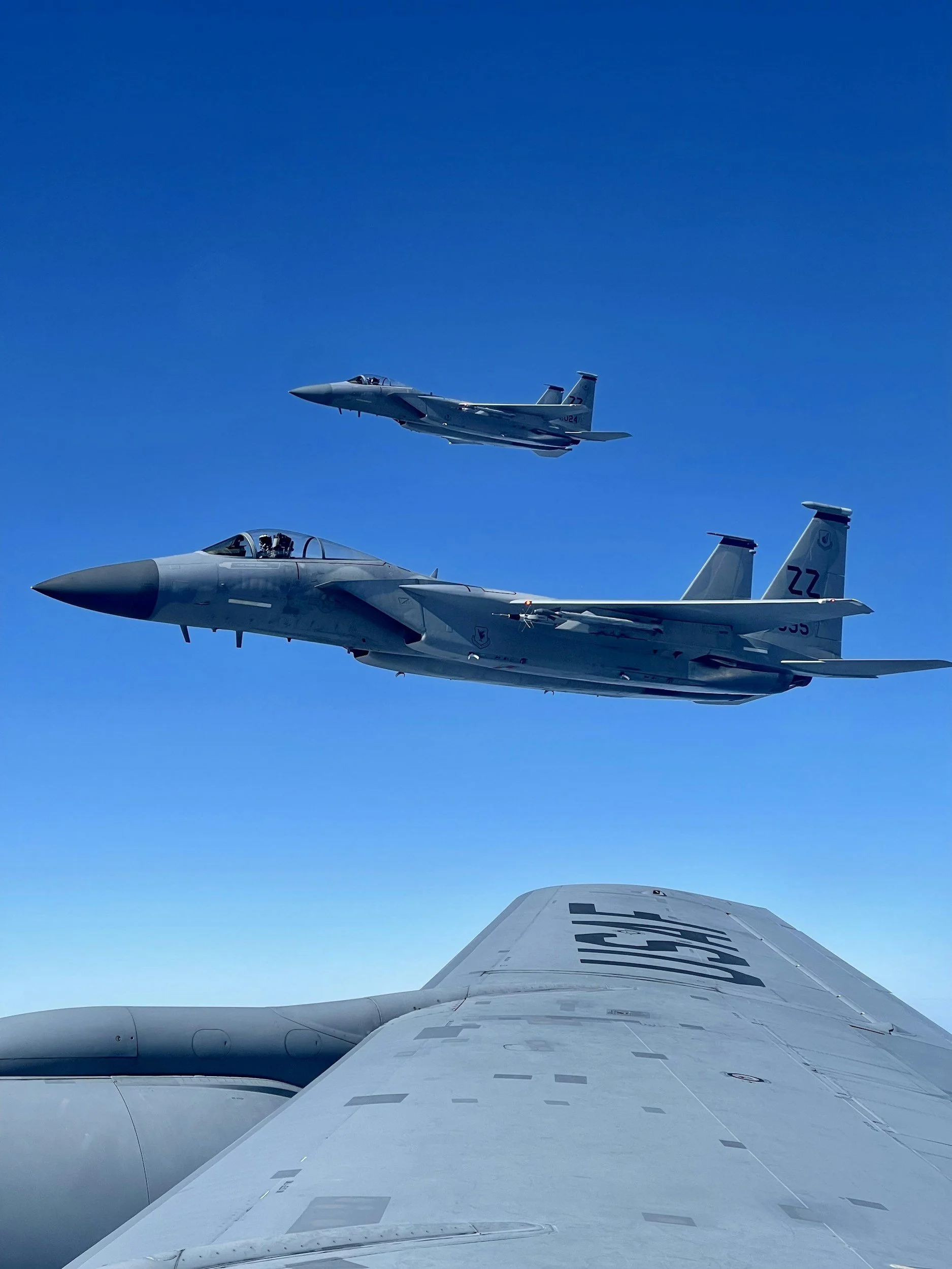 Two fighter jets flying in a clear blue sky, seen from a plane wing below.