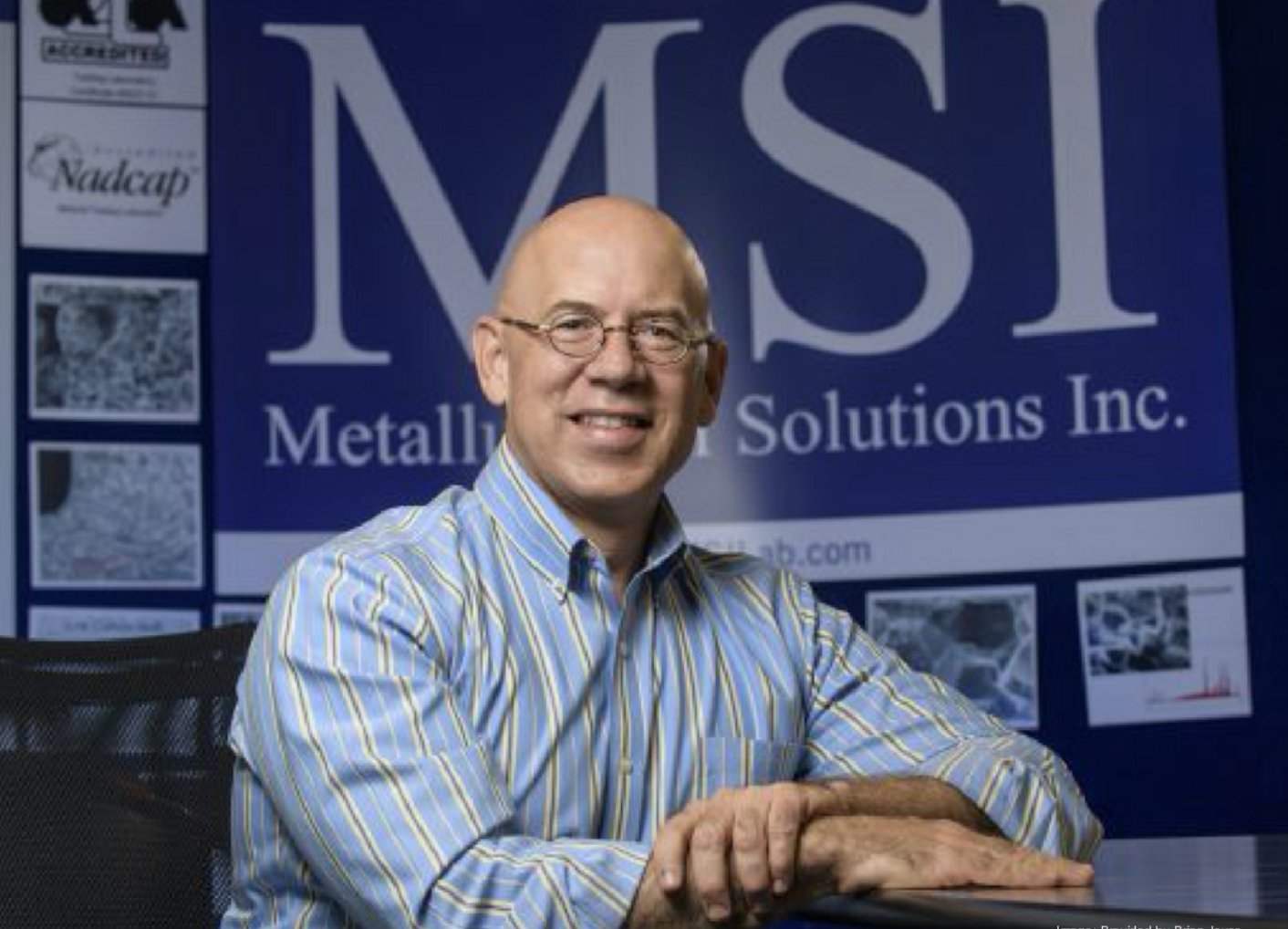 A smiling man with glasses and a striped shirt sitting at a desk in front of a large blue banner that reads 'Metallurgical Solutions Inc.' with smaller images and text on the side.