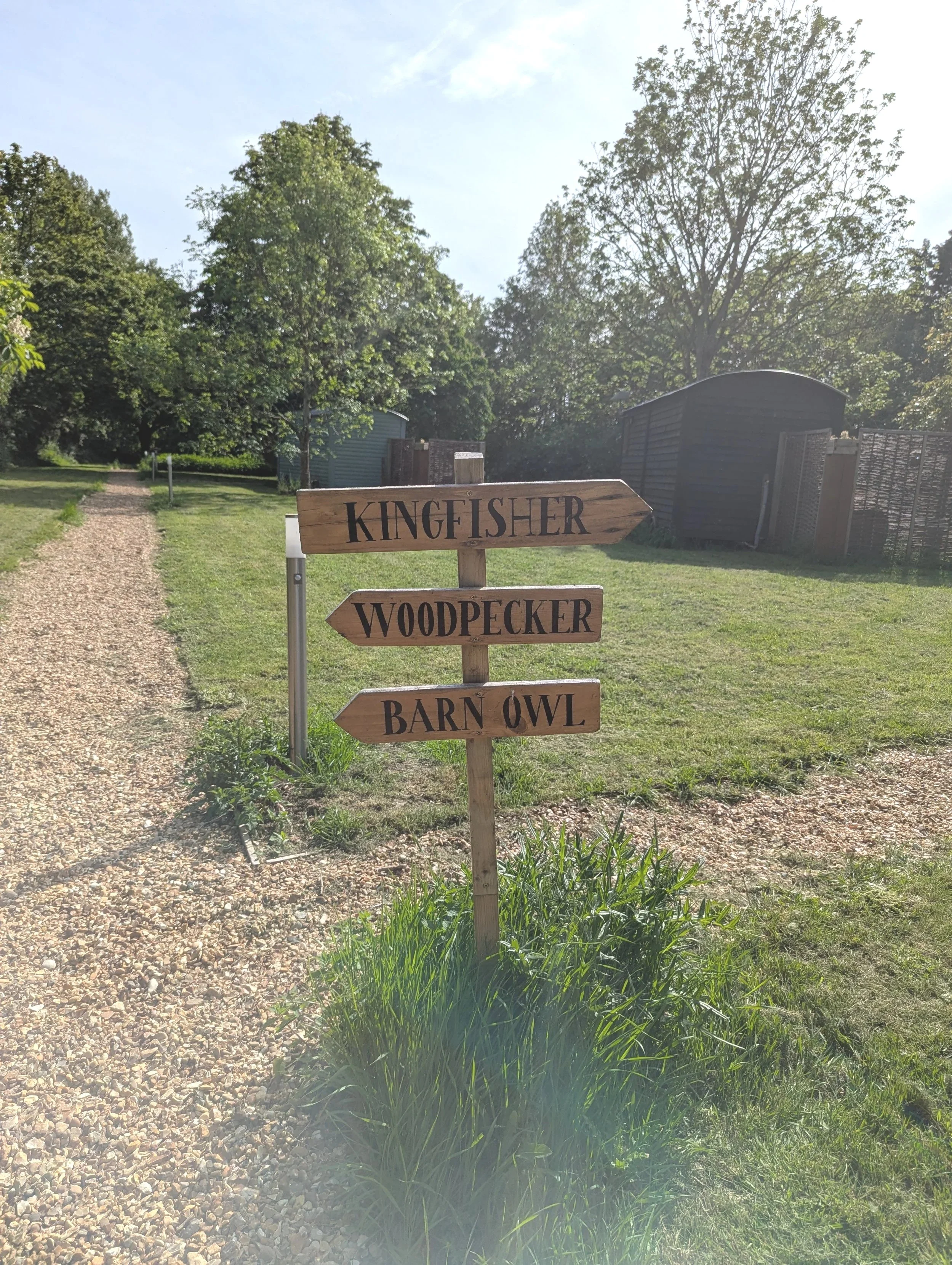 A wooden signpost with three arrows pointing right, with words 'KINGFISHER,' 'WOODPECKER,' and 'BARN OWL' written on them, located in a grassy outdoor area with trees and a gravel pathway.