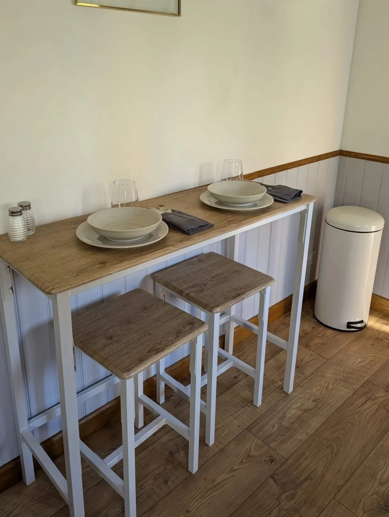 A small dining area with a wooden high table set with two large bowls, two glasses, gray napkins, and salt and pepper shakers. There are three wooden stools with white frames, a trash can, and a section of a white-paneled wall with wood trim.