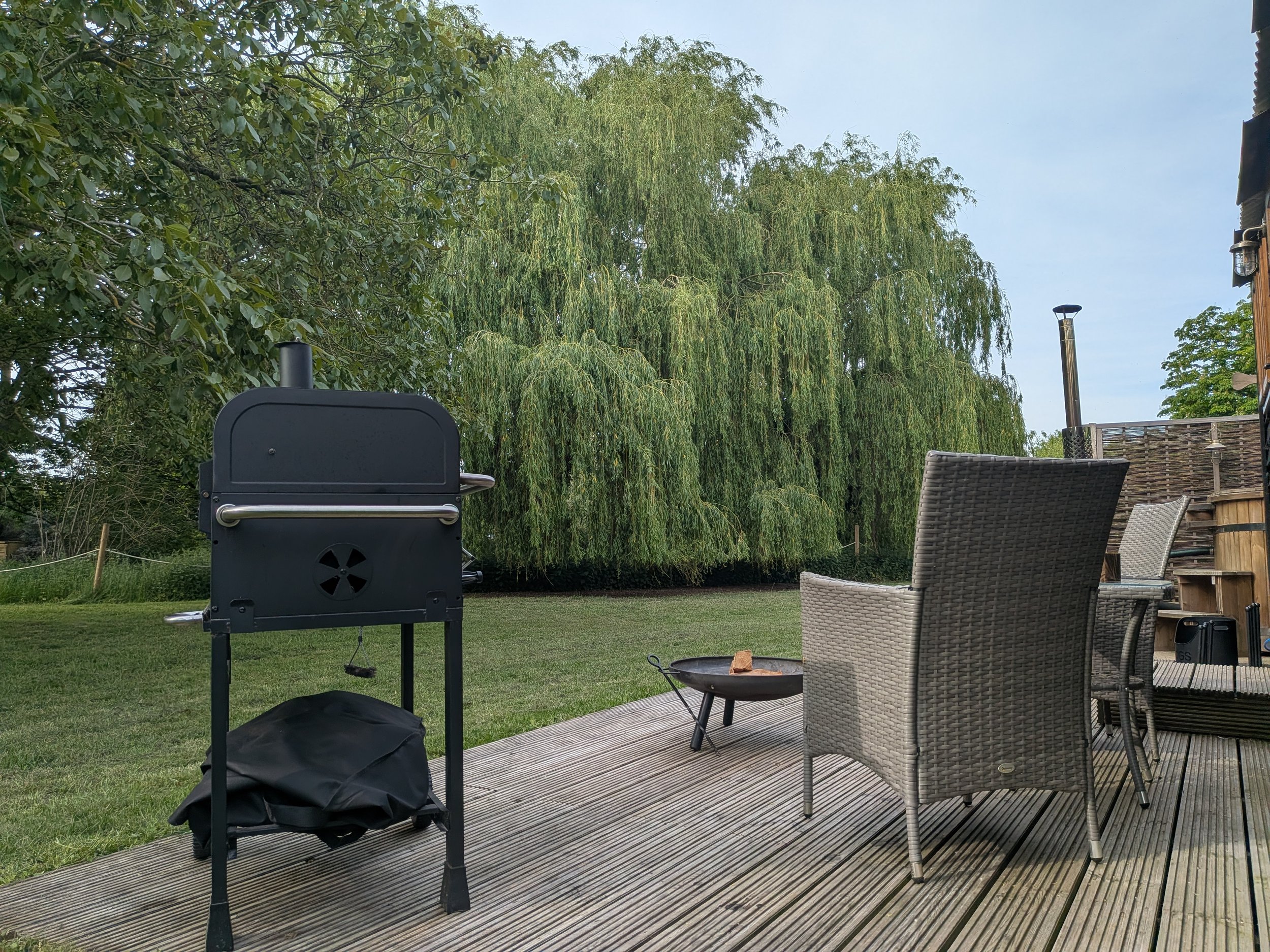Backyard patio with outdoor grill, wicker chairs, a small fire pit, and lush green trees in the background.