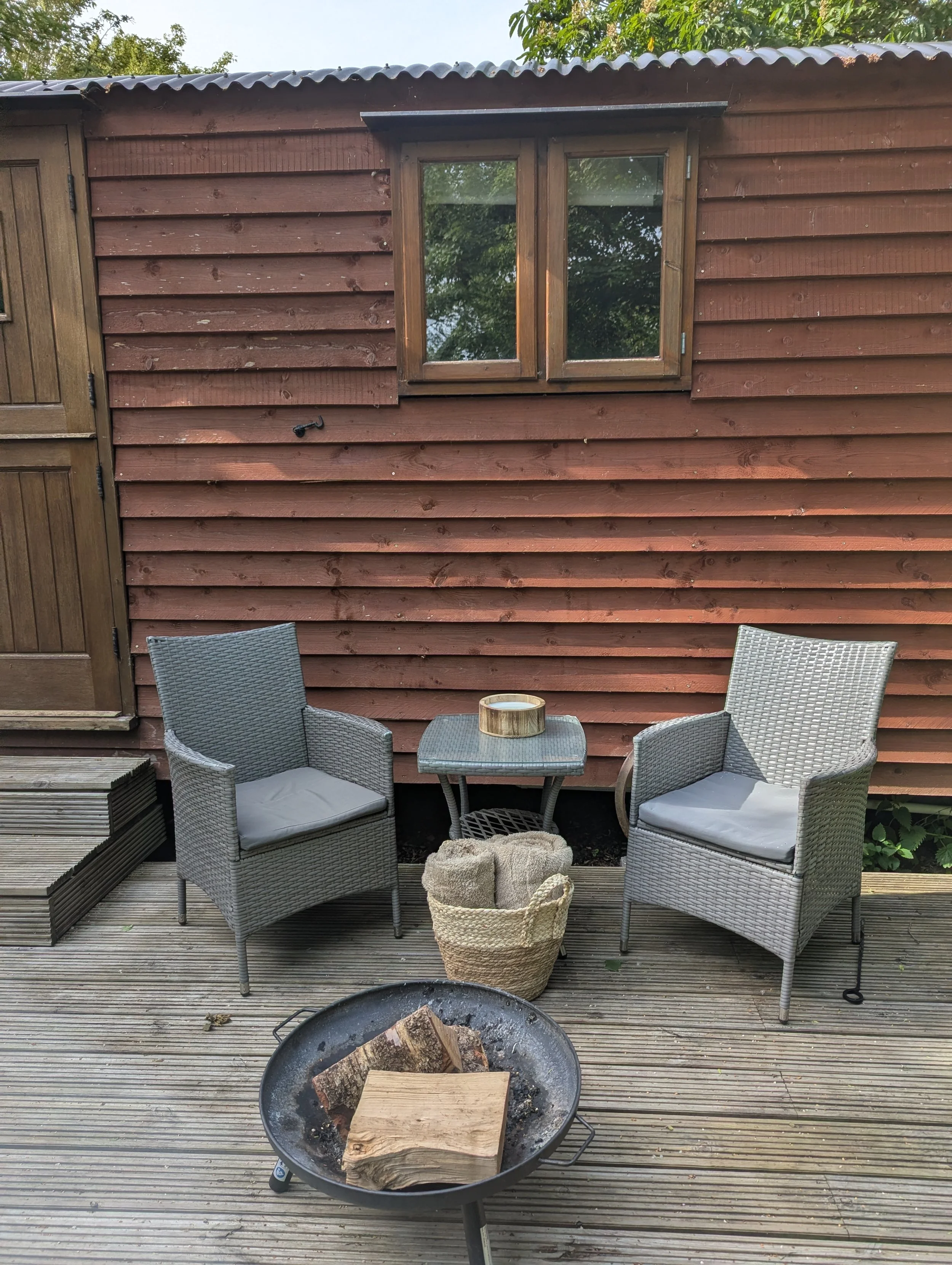 A wooden deck with three gray outdoor chairs and a small matching table with a ceramic bowl, in front of a shepherds hut with brown horizontal siding and a window. There is a fire pit with wood pieces in the foreground.