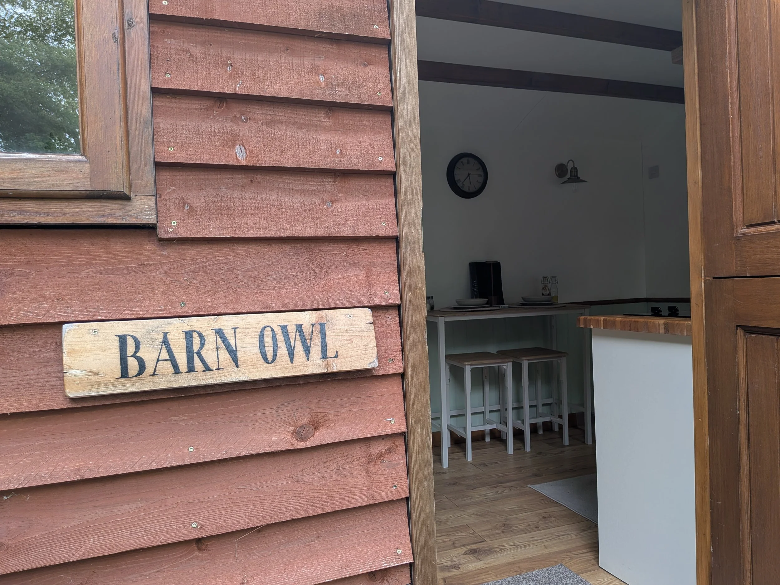 Open hut door revealing a modern interior with white walls, a clock, a wall-mounted light fixture, and a table with chairs.
