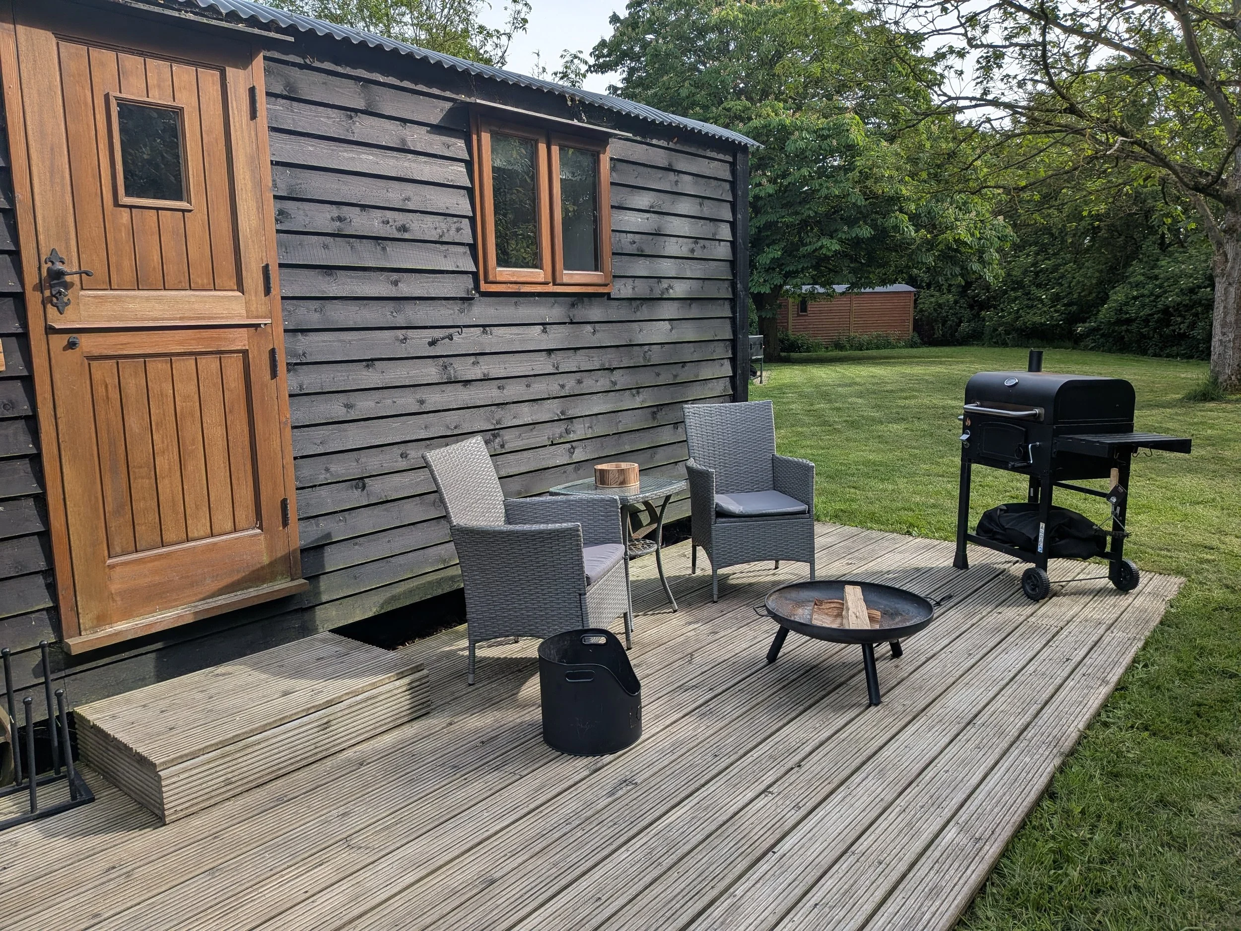 A private patio with wooden decking, outdoor seating, a black grill, a fire pit, and a wooden shepherds hut, surrounded by green trees and grass.