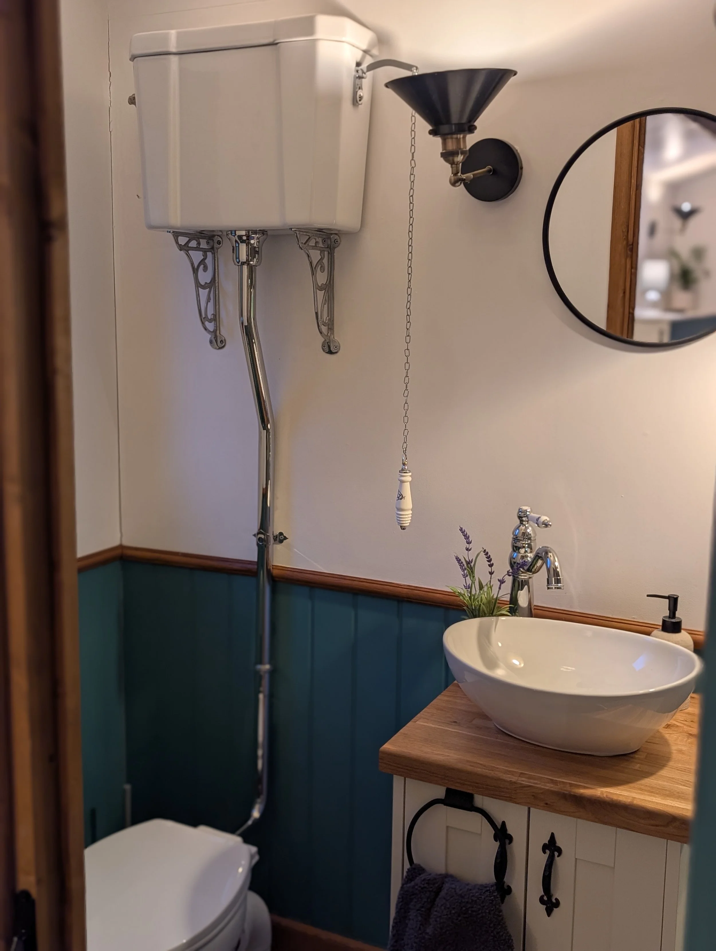 An ensuite bathroom corner with a vintage toilet with a high tank, a black wall-mounted sink faucet, a round mirror, a small plant on the sink counter, and a white vessel sink on a wooden countertop.