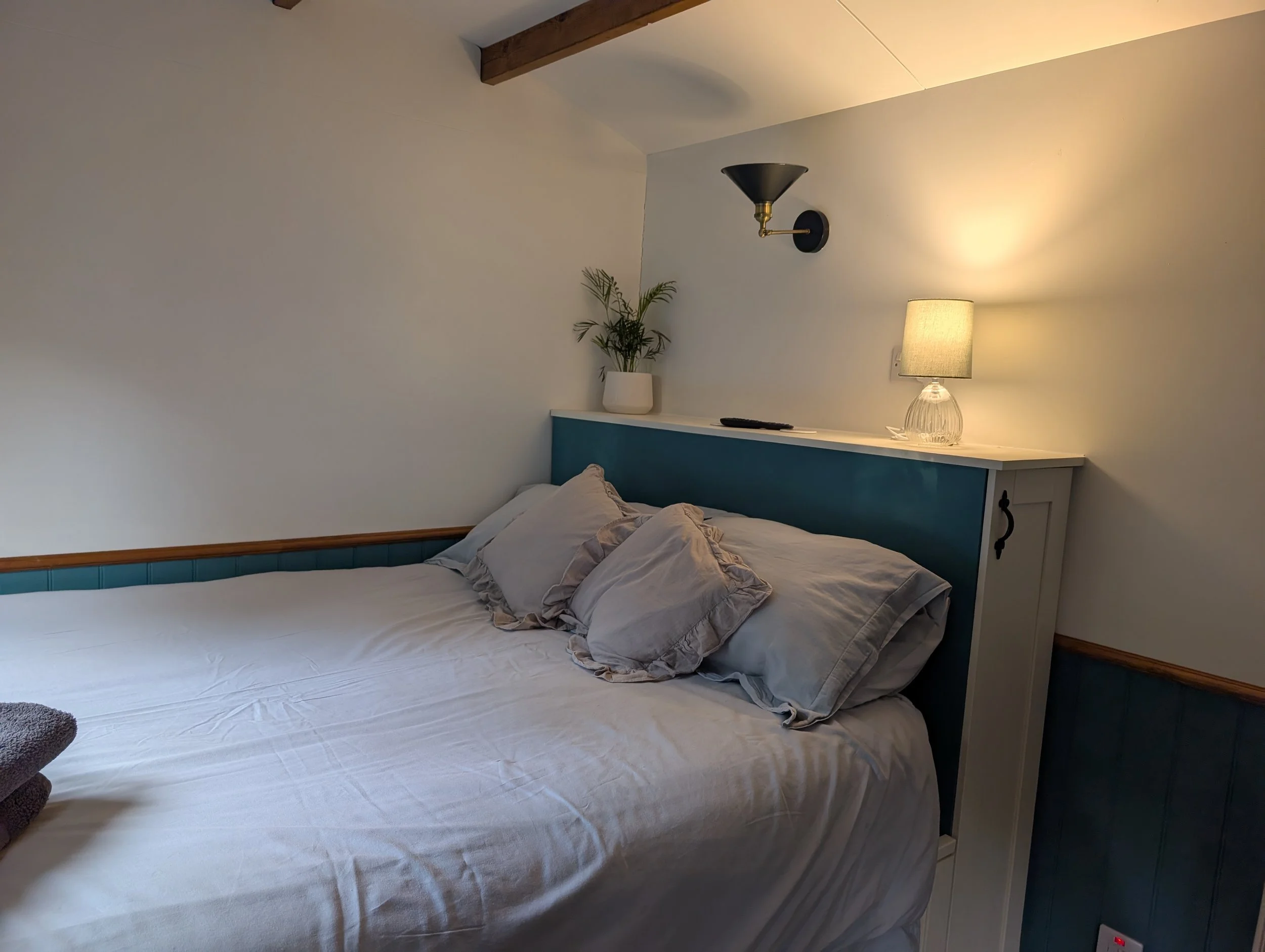 A king-sized bed, pillows, a nightstand with a lamp and a potted plant, a wall with a wall sconce, and a wooden beam on the ceiling.