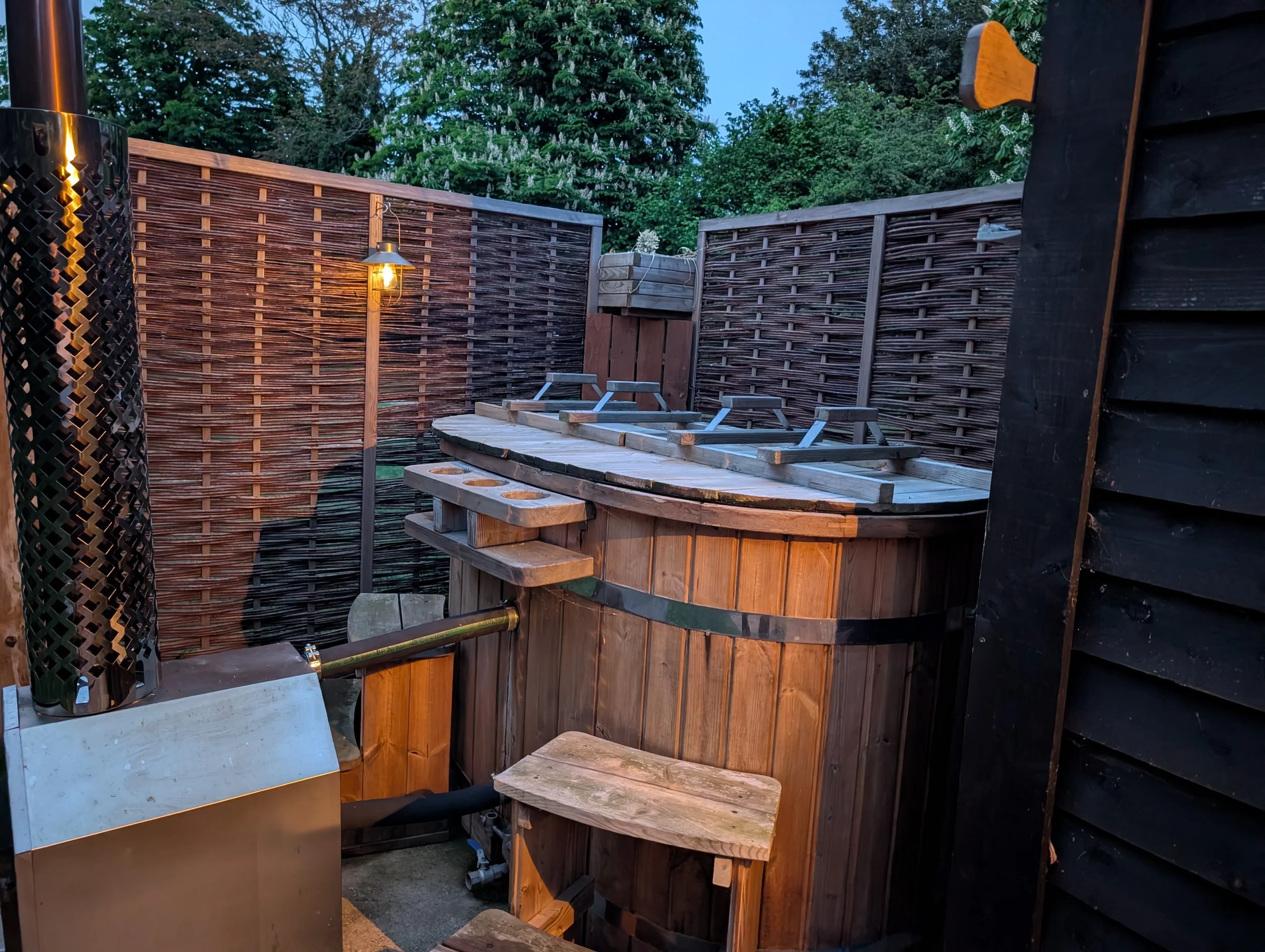 An outdoor hot tub surrounded by wooden privacy fences, with a light fixture on one of the fences and a potted plant on a shelf. Trees and a blue sky are visible in the background.