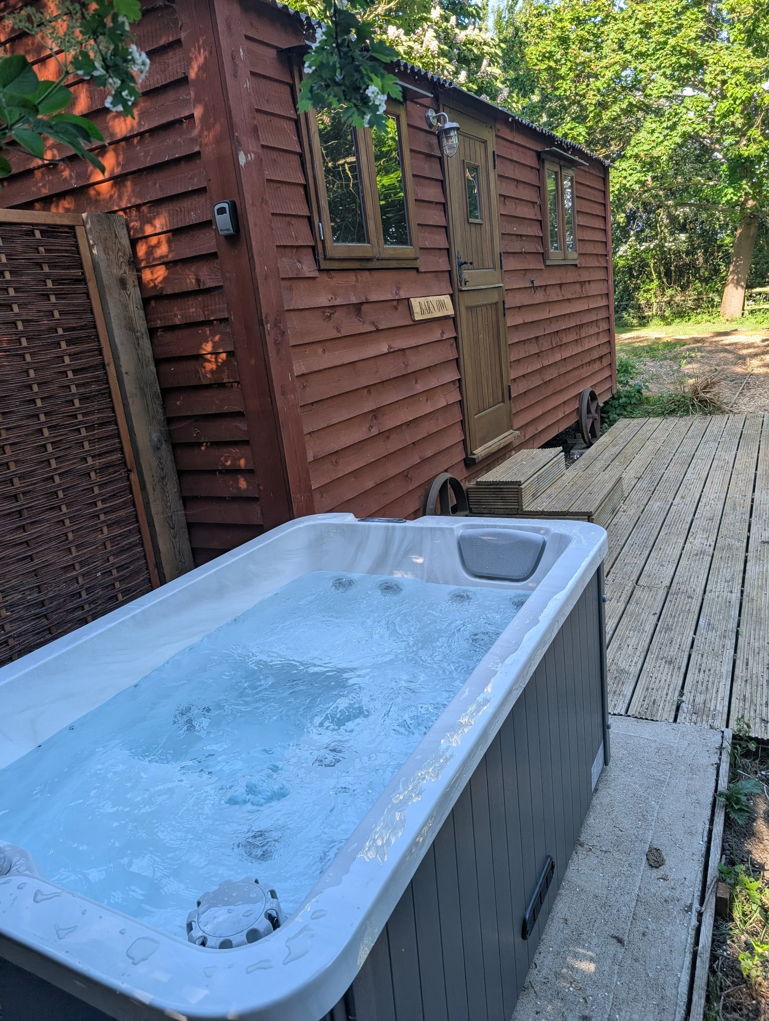 An outdoor hot tub filled with water on a wooden deck, with a shepherds hut with a wooden door and windows in the background, surrounded by trees and greenery.