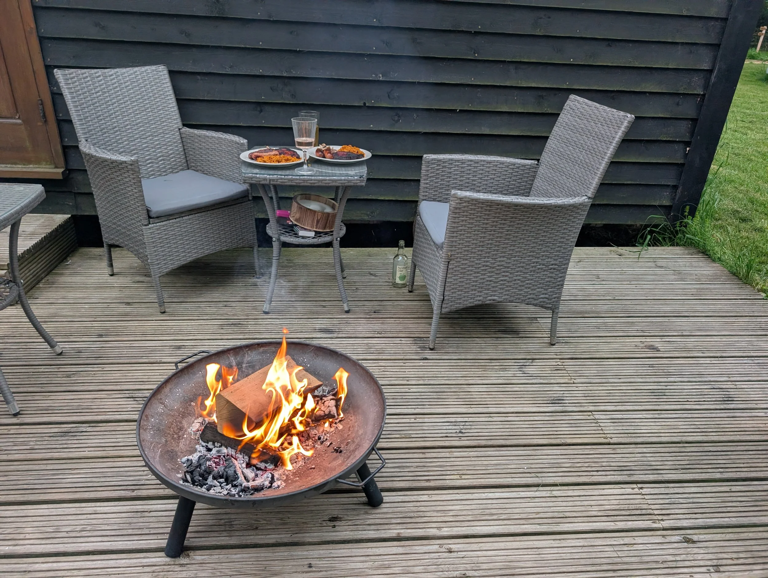 A backyard deck with a fire pit in the foreground and two wicker chairs with cushions and a small table with plates of food and drinks against a black shepherd's hut.