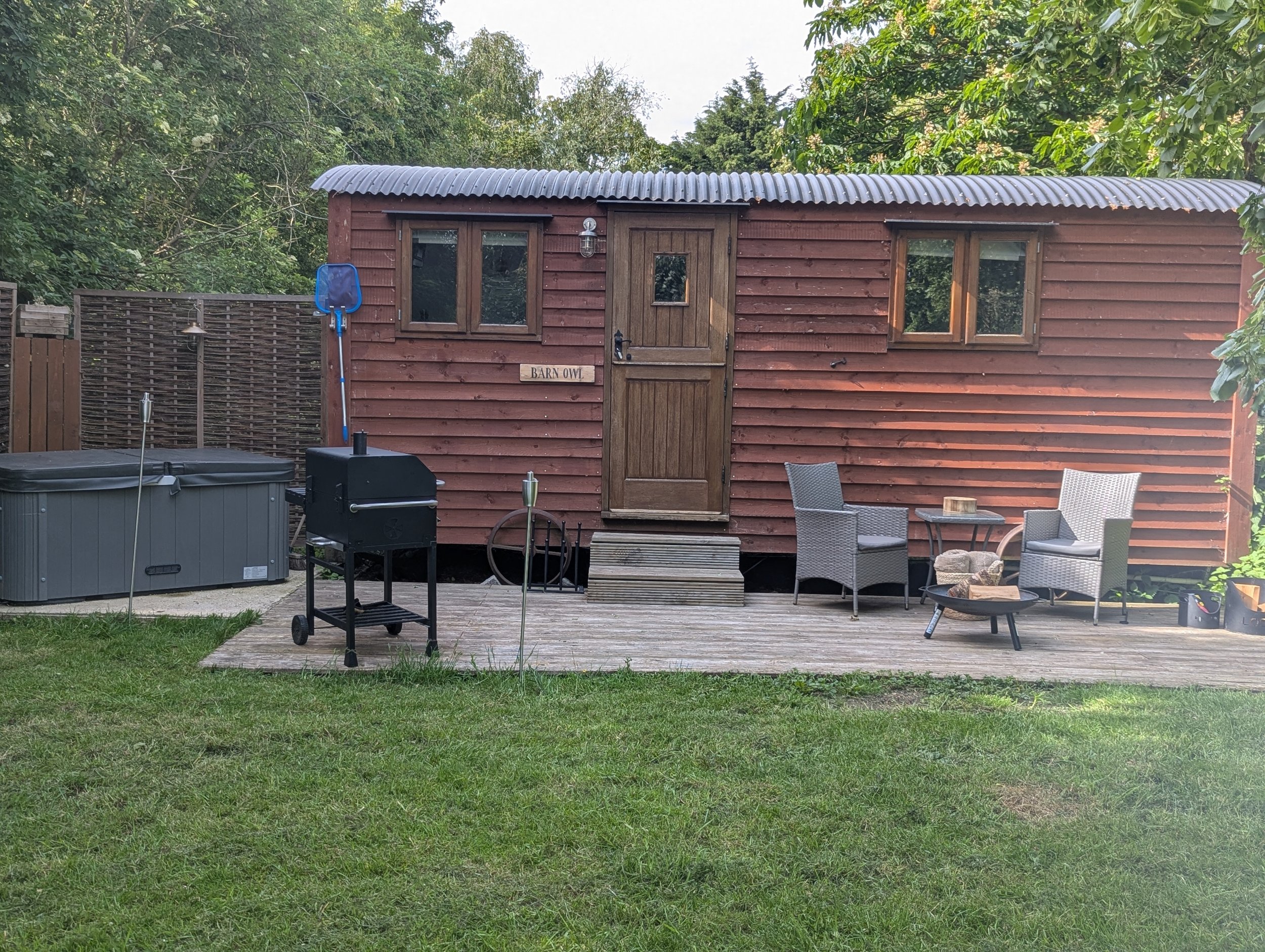 A private shepherds hut, surrounded by green trees, with outdoor furniture, a barbecue grill, and a small garden area in the yard.